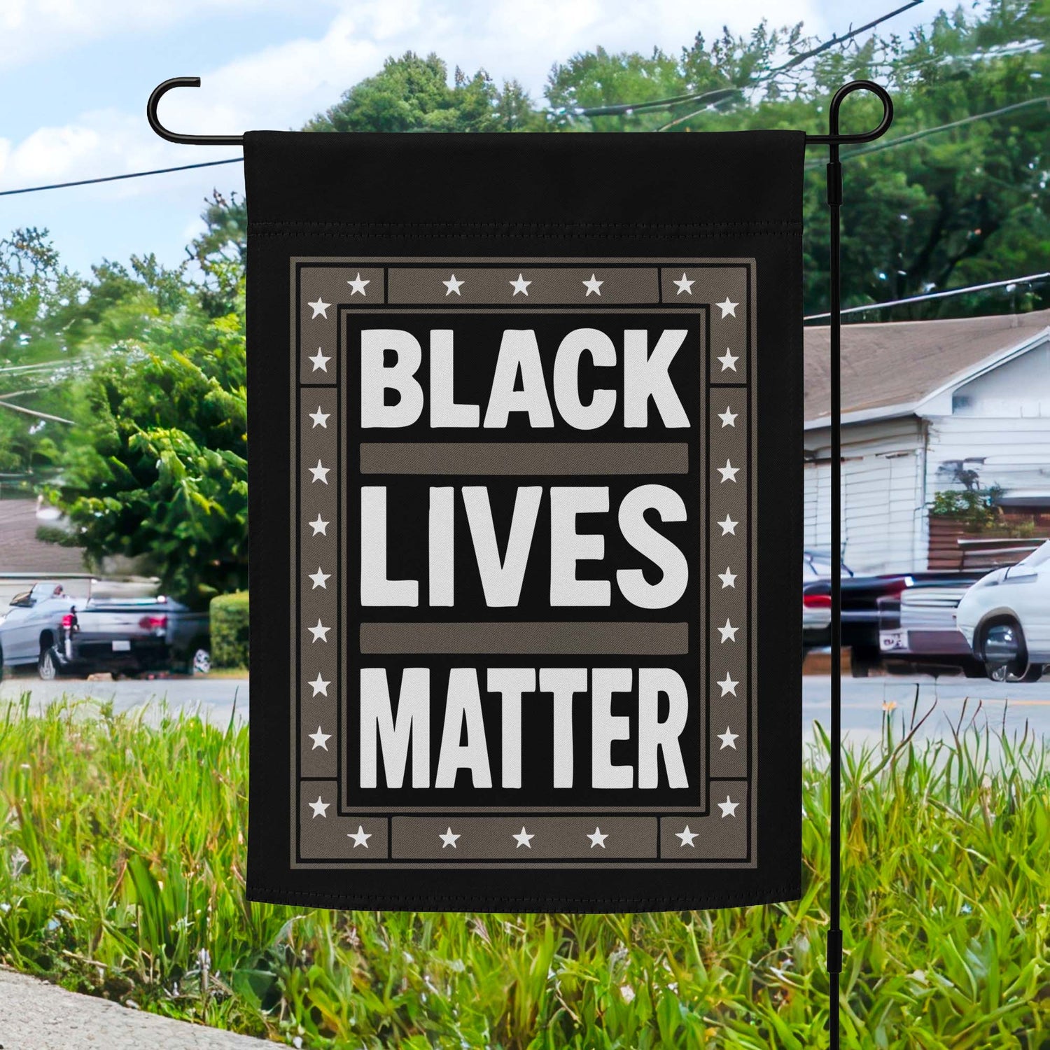 Black Lives Matter garden flag with bold white text on black background and star border, displayed outdoors on a flagpole in front of a home.