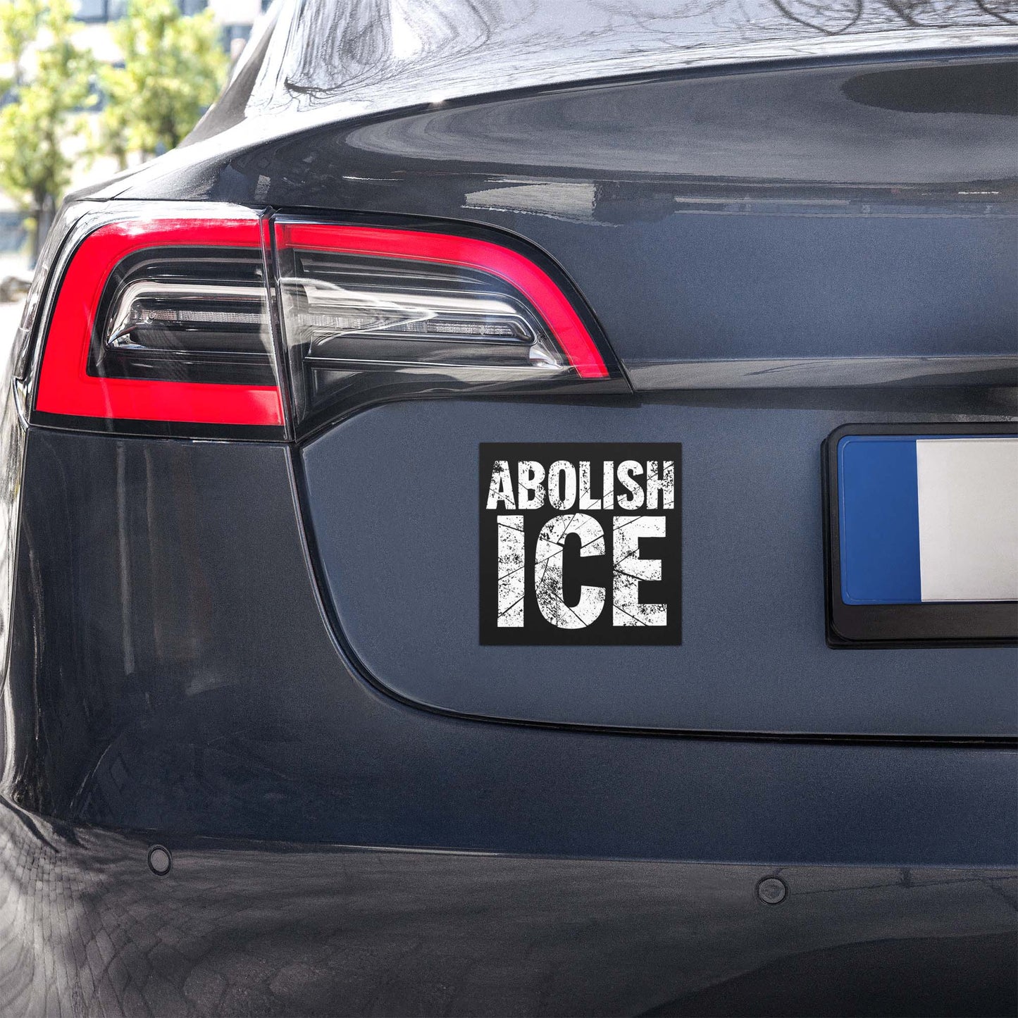 Car magnet mounted on vehicle bumper showing weathered block typography with cracked texture, high-contrast black and white design, printed message “ABOLISH ICE.”