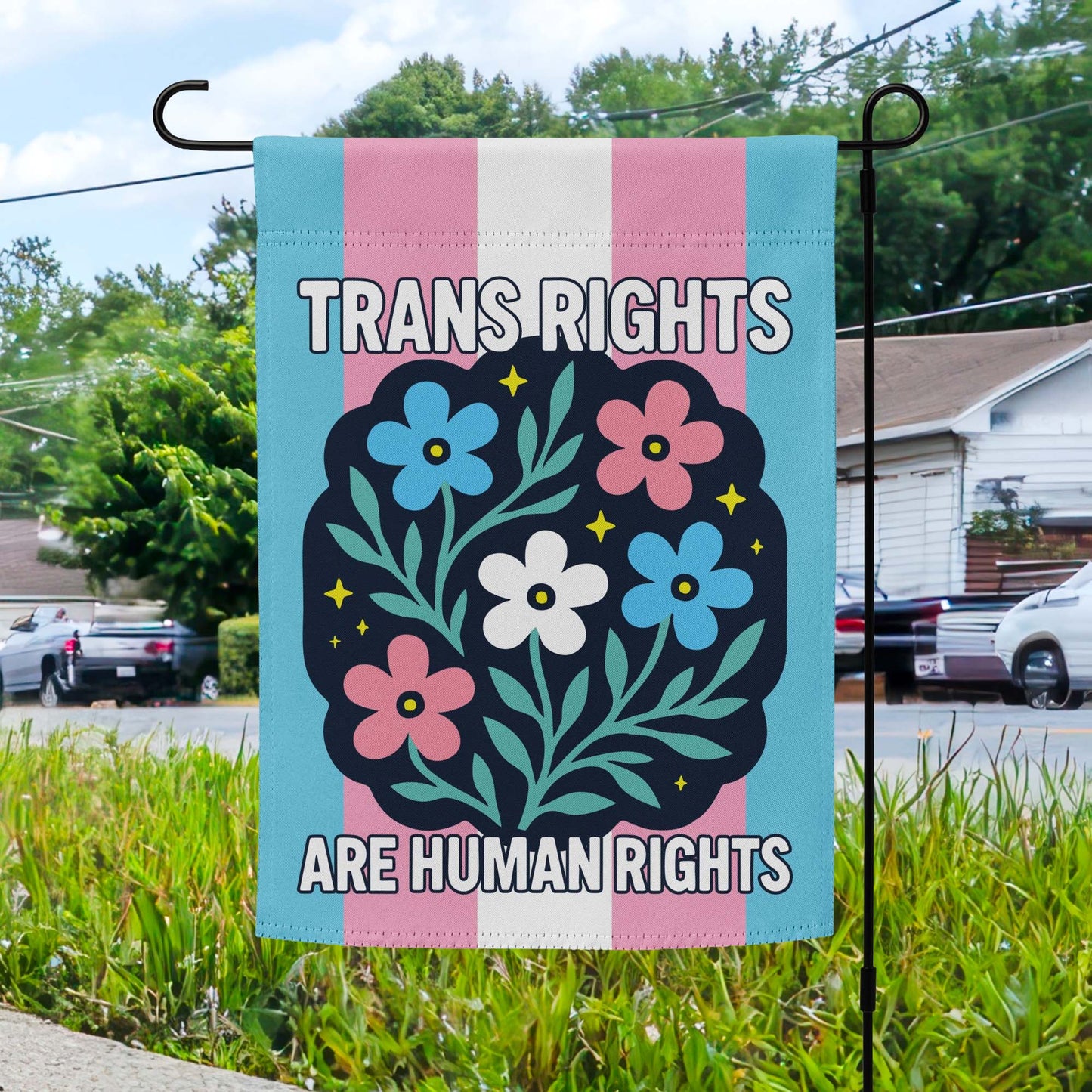 A garden flag with transgender flag colors and a central floral design, displaying "TRANS RIGHTS ARE HUMAN RIGHTS" in bold lettering, set against the background of a suburban yard.