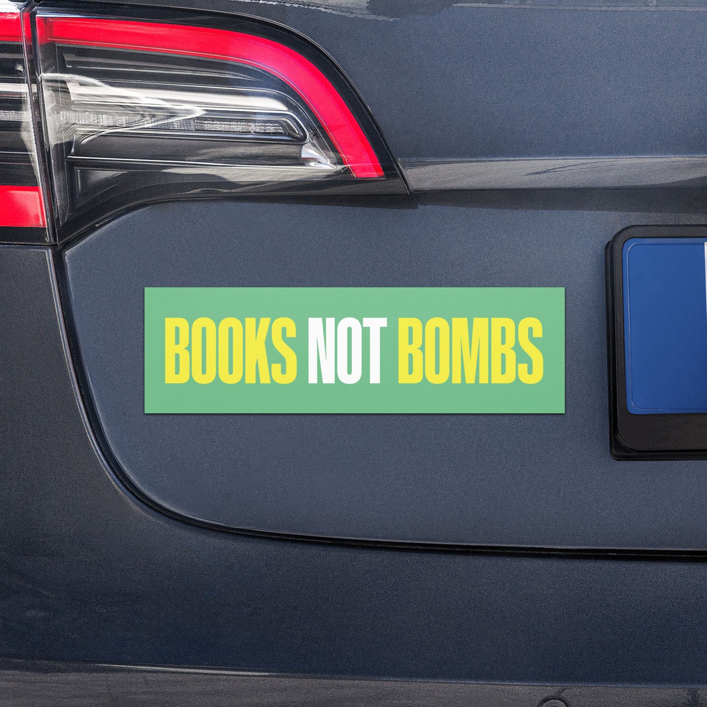 Car magnet displayed on vehicle bumper with bold phrase “BOOKS NOT BOMBS” in yellow and white lettering on green background.