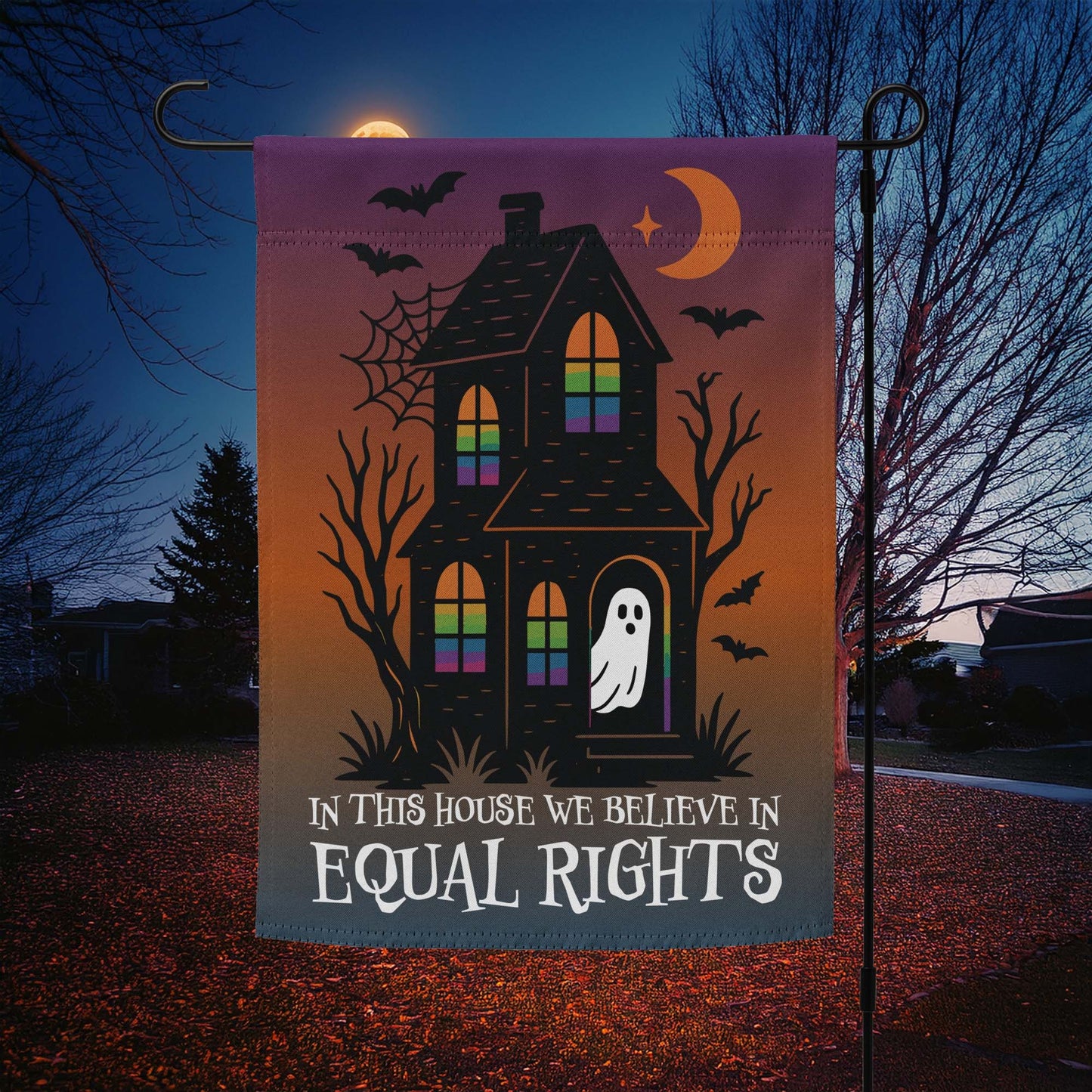 “In This House We Believe in Equal Rights” Halloween garden flag with haunted house, ghost, bats, and crescent moon, displayed outdoors at night.