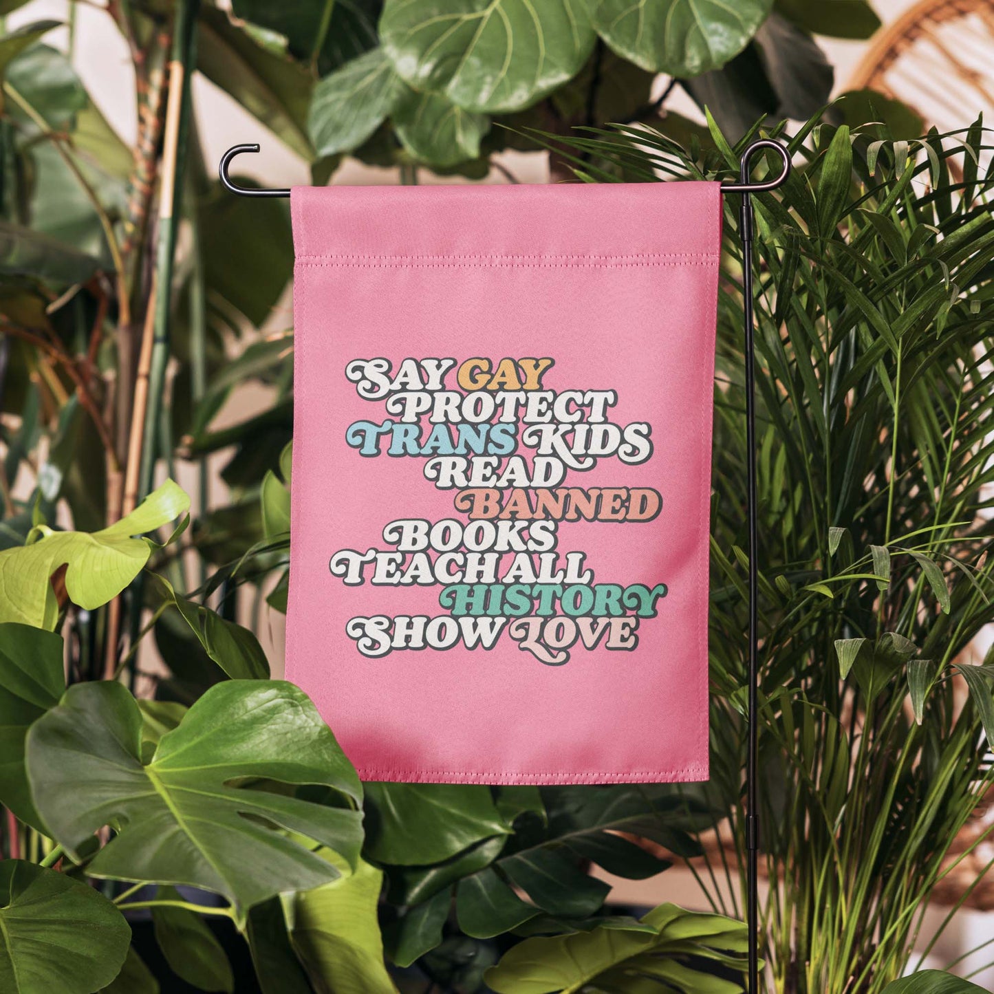 Pink garden flag displayed among green plants featuring bold retro stacked typography reading “Say Gay Protect Trans Kids Read Banned Books Teach All History Show Love.”