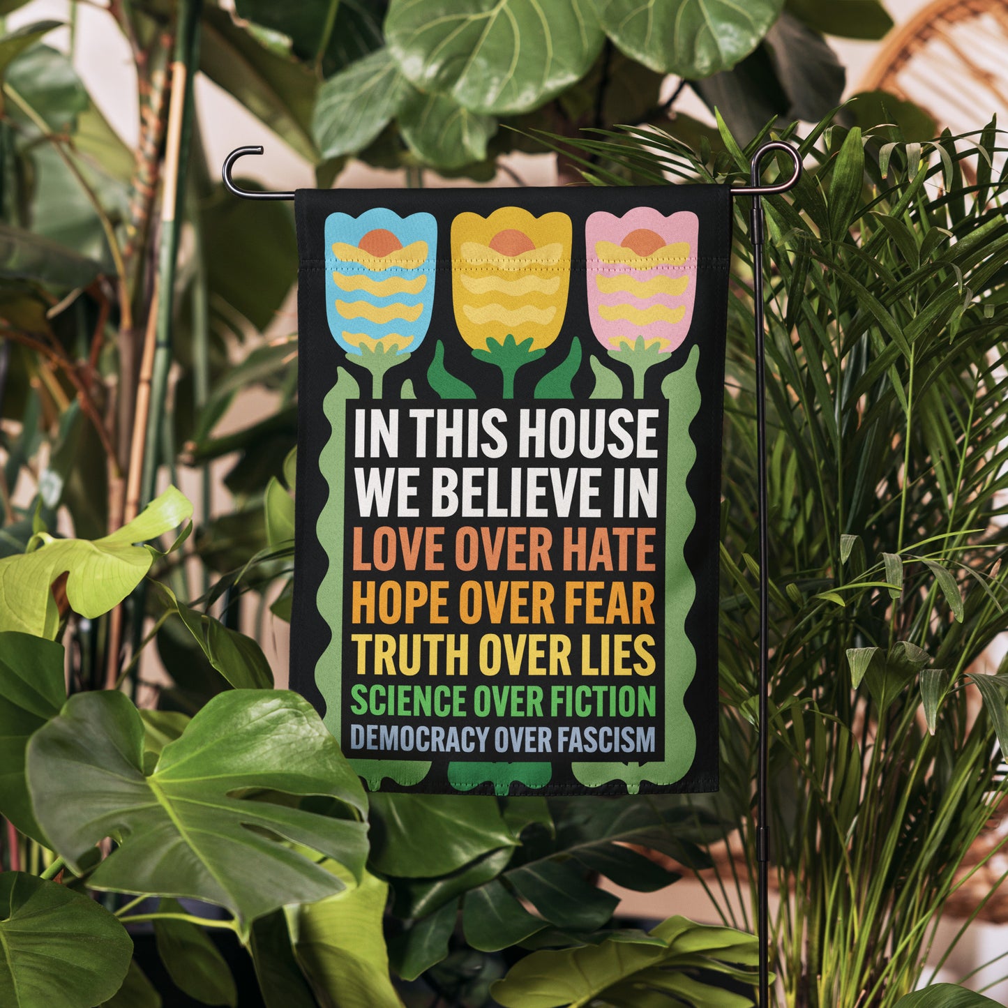 Garden flag reading “In This House We Believe In Love Over Hate, Hope Over Fear, Truth Over Lies, Science Over Fiction, Democracy Over Fascism” shown hanging in greenery.