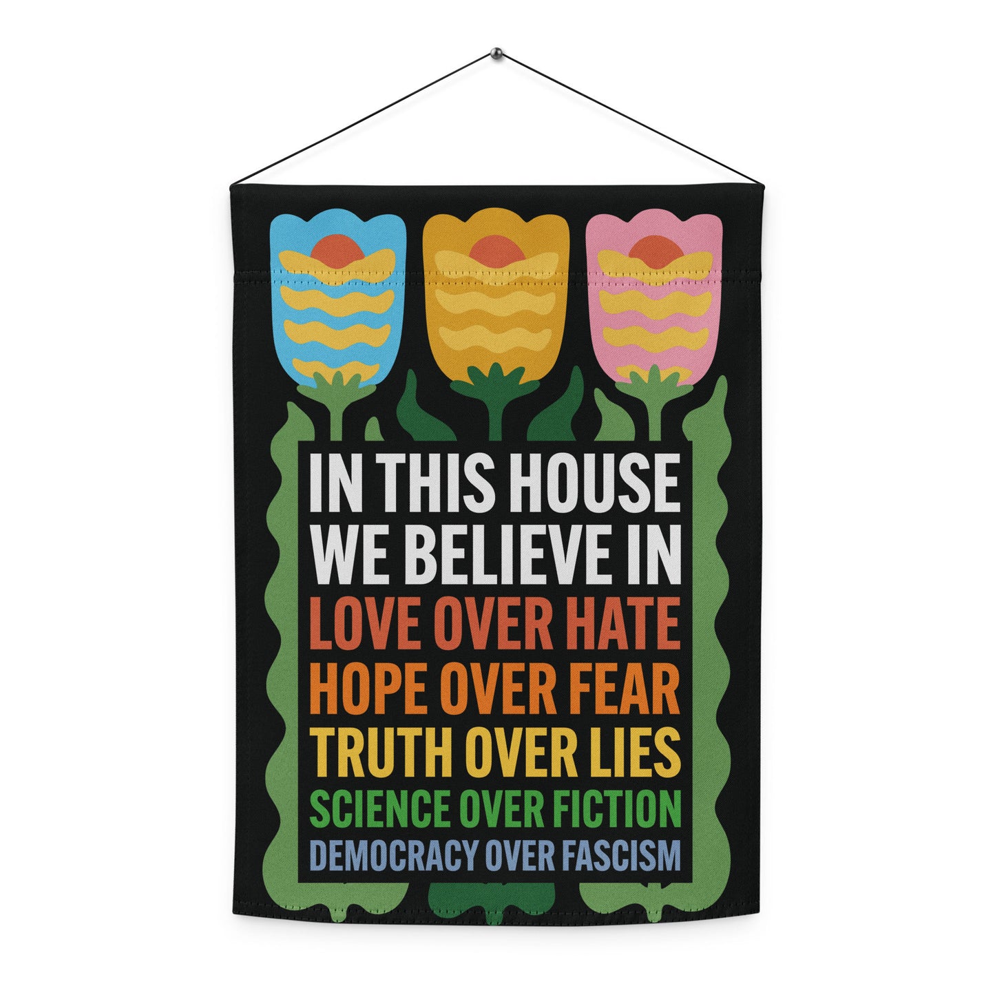 Front view of the “In This House We Believe” tulip garden flag with rainbow-colored text and floral top design.