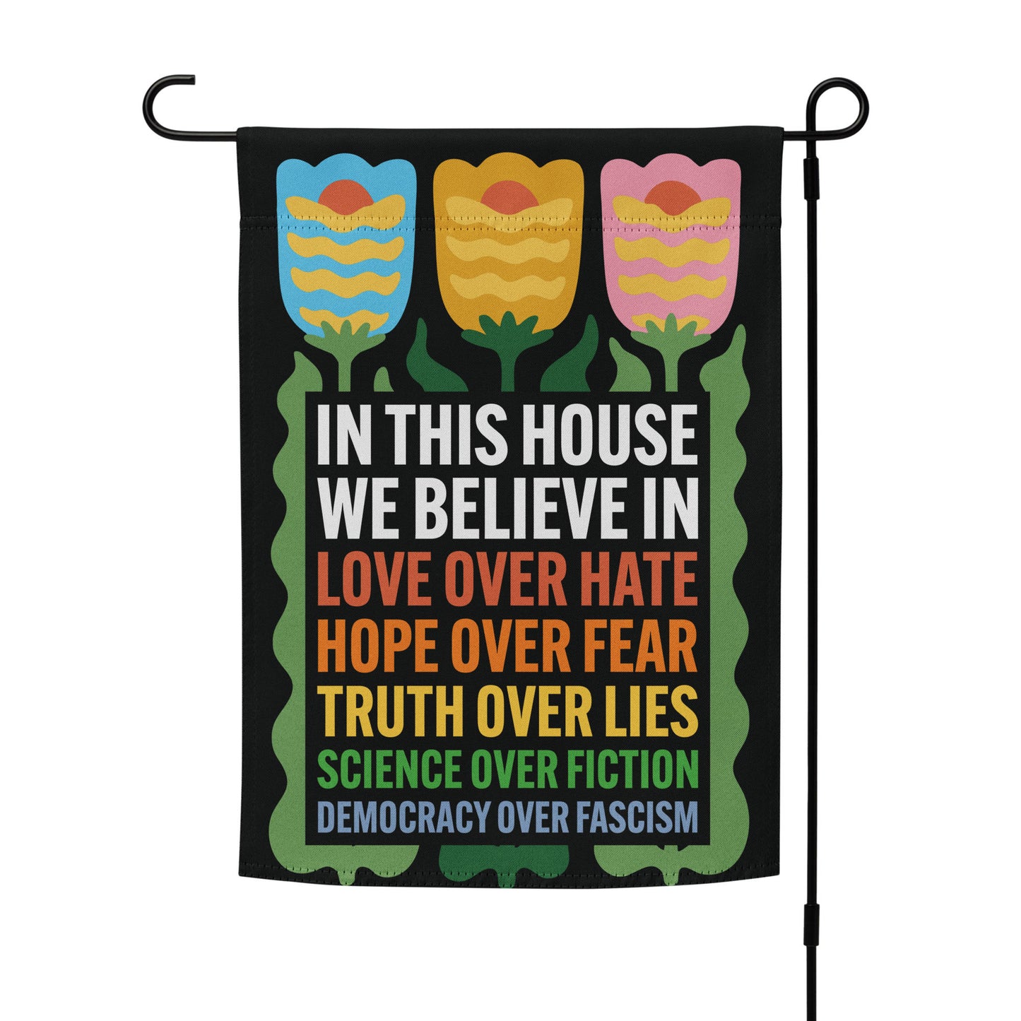 “In This House We Believe” progressive garden flag featuring tulip flowers and bold rainbow text, displayed on a flag stand.