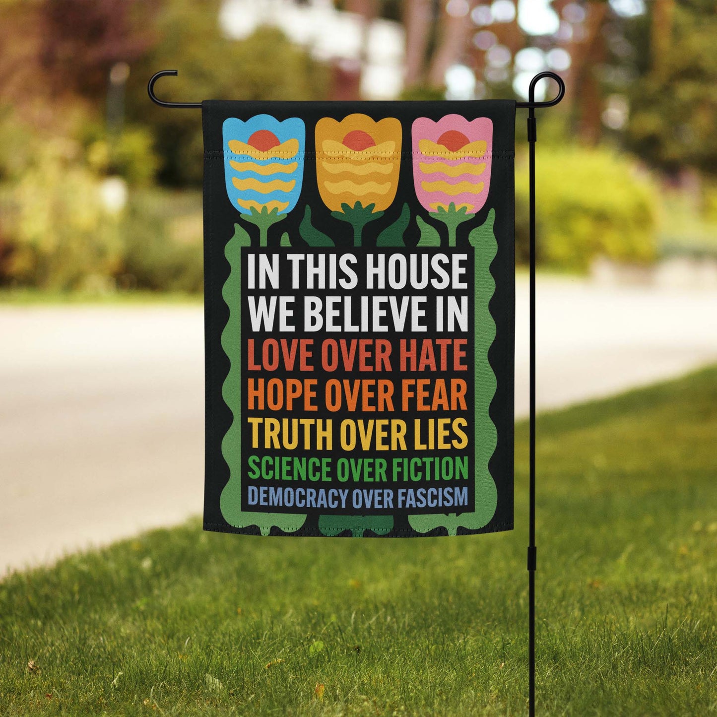 Progressive “In This House We Believe” garden flag with tulip design and rainbow text displayed on a flag stand in a grassy yard.
