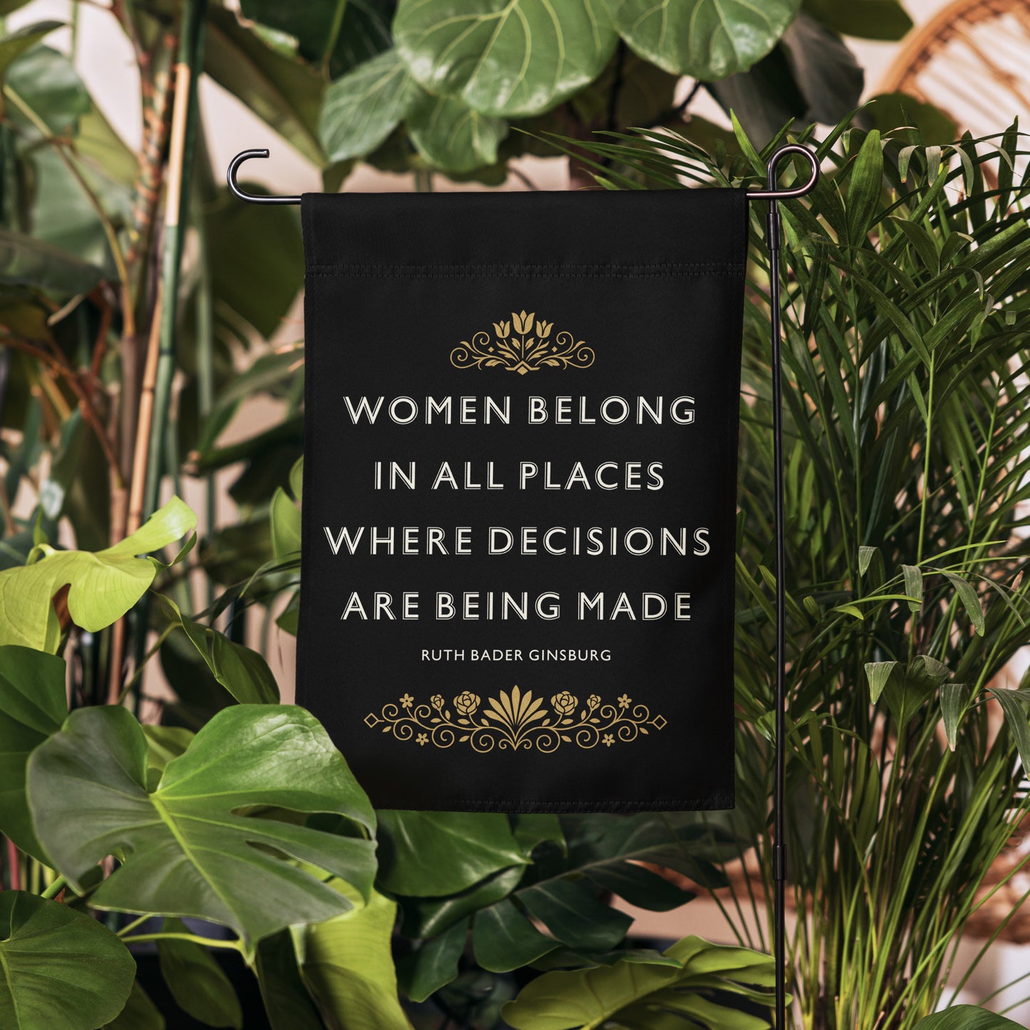 “Women Belong In All Places Where Decisions Are Being Made” Ruth Bader Ginsburg garden flag displayed in greenery, with gold Art Deco floral details on black background.