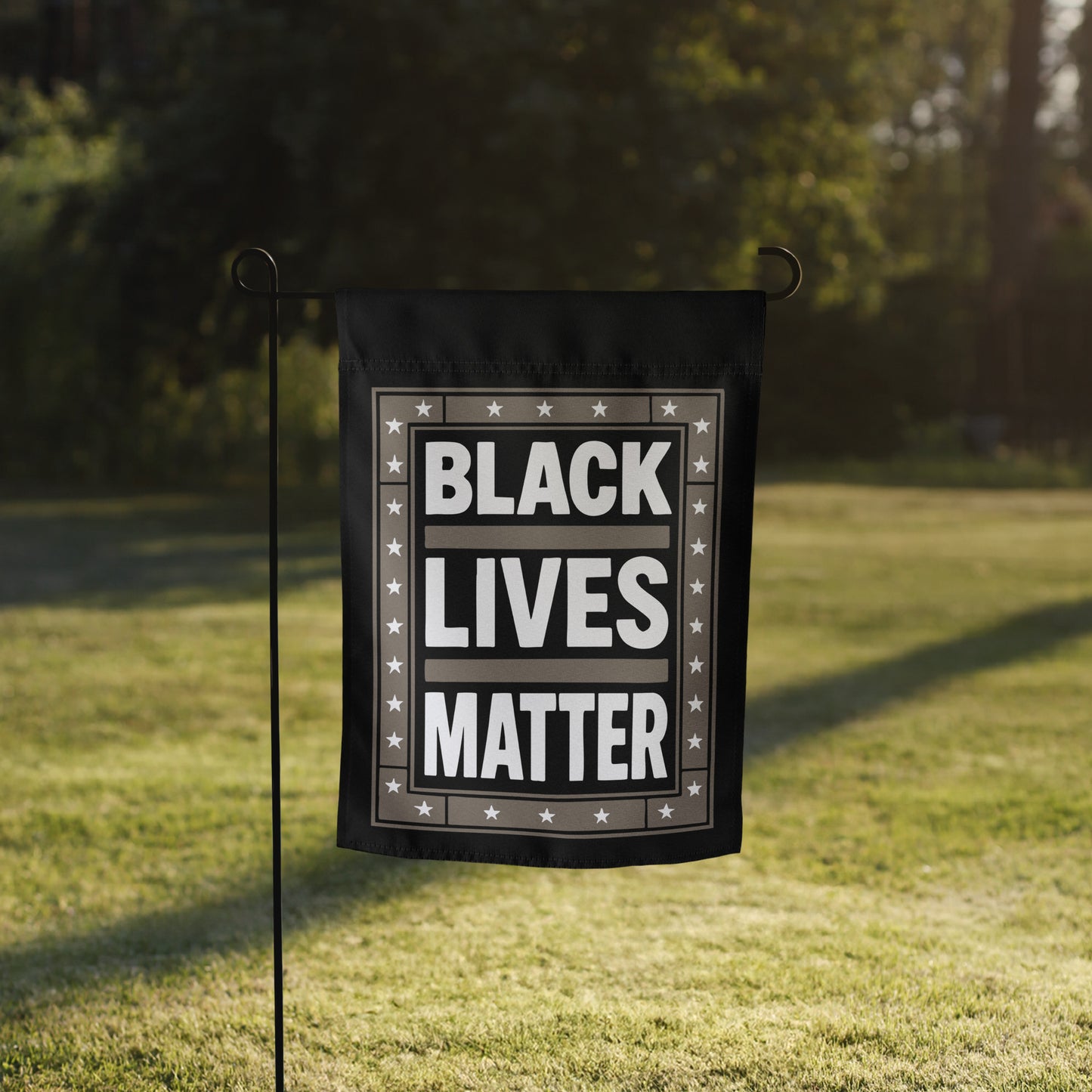 Black Lives Matter garden flag with star border shown on a flag stand in a grassy yard with sunlight behind it.