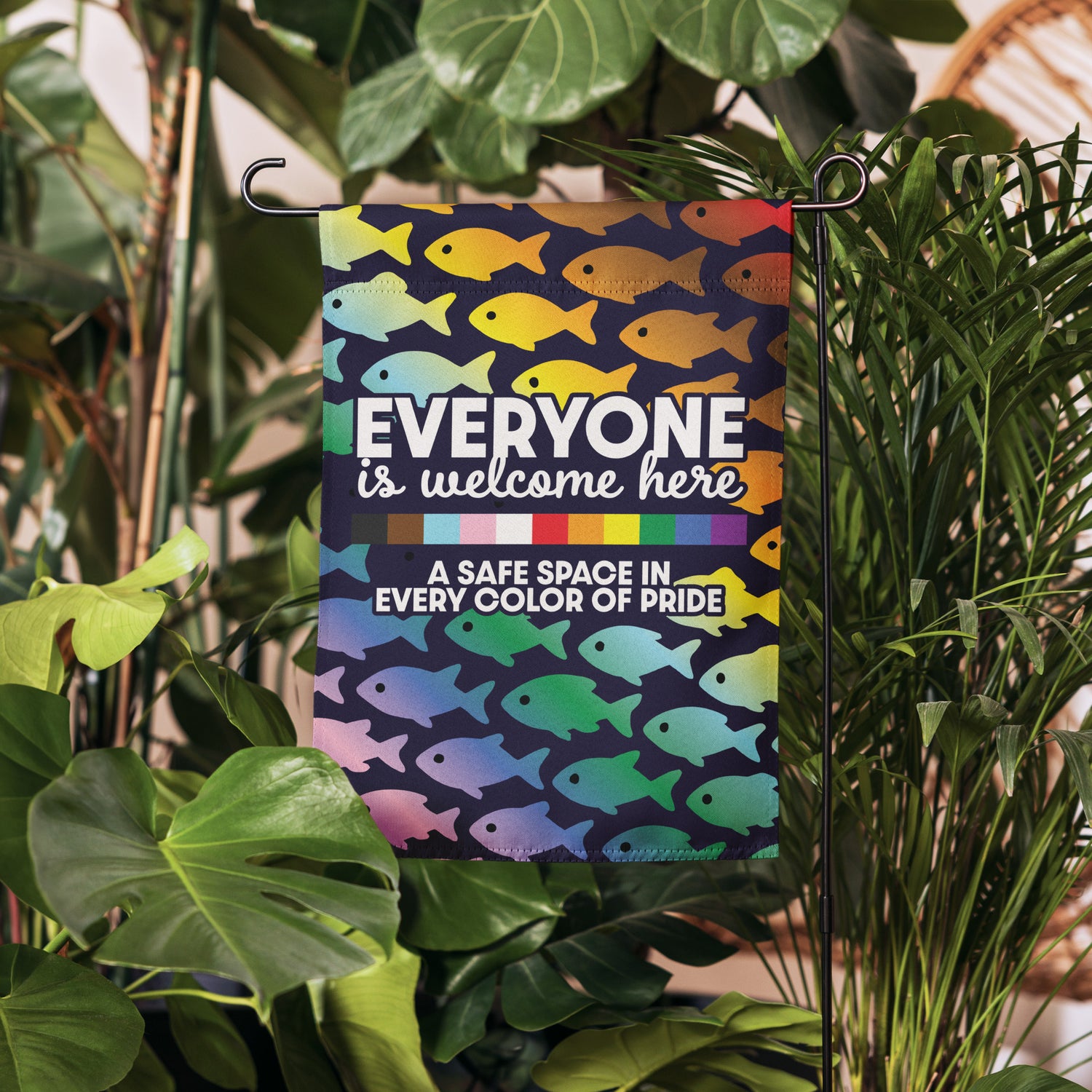 Inclusive pride garden flag reading “Everyone Is Welcome Here – A Safe Space In Every Color of Pride” shown hanging among greenery.