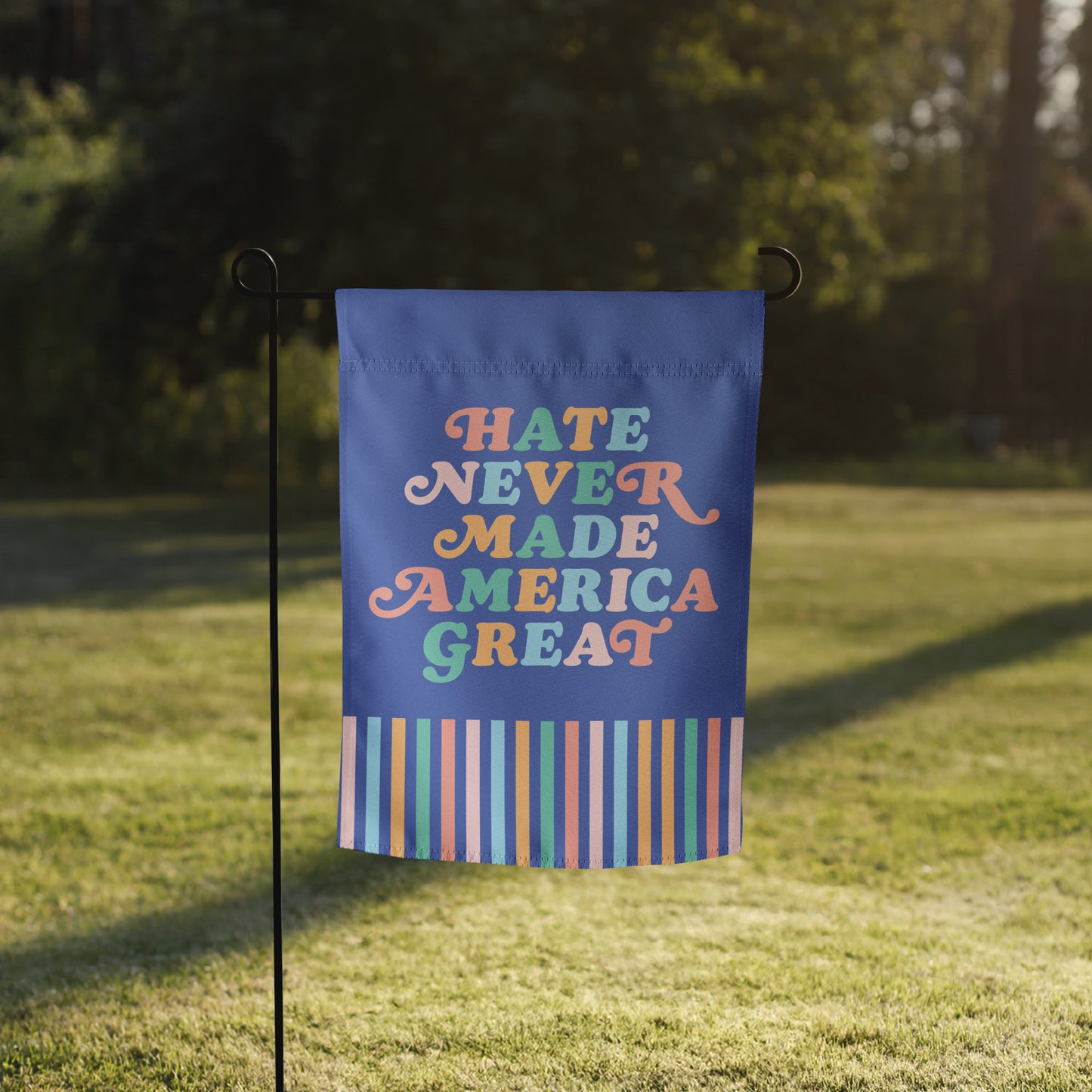 Progressive garden flag reading “Hate Never Made America Great” with colorful stripes at the bottom, shown on a flag stand in a grassy yard.