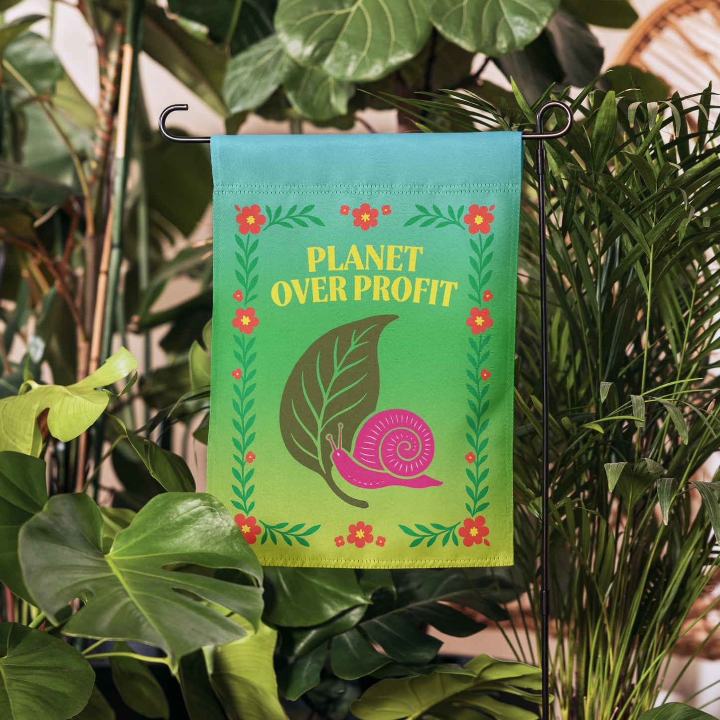 Progressive climate justice garden flag with snail and leaf artwork, “Planet Over Profit” slogan, shown hanging among greenery.