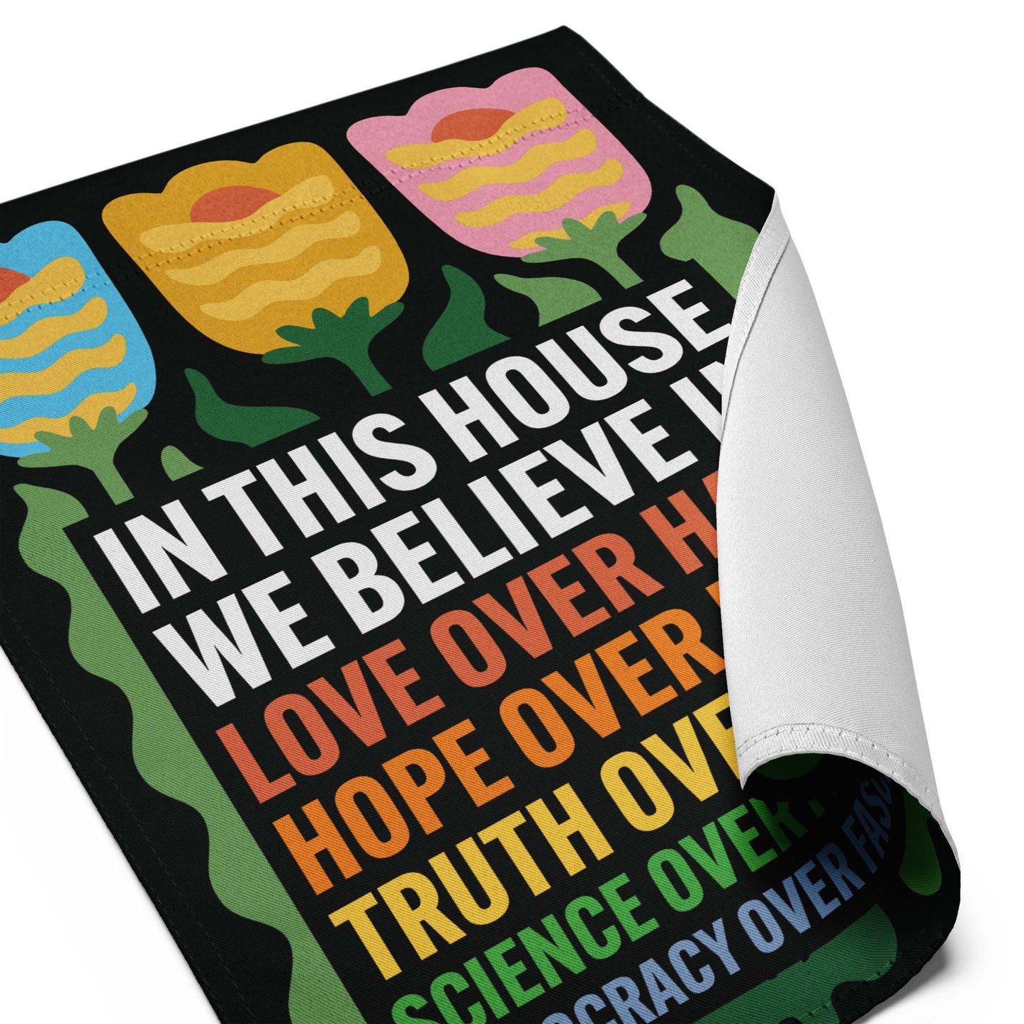 Close-up of the “In This House We Believe” tulip garden flag with curled fabric edge, showing colorful flowers and bold lettering.