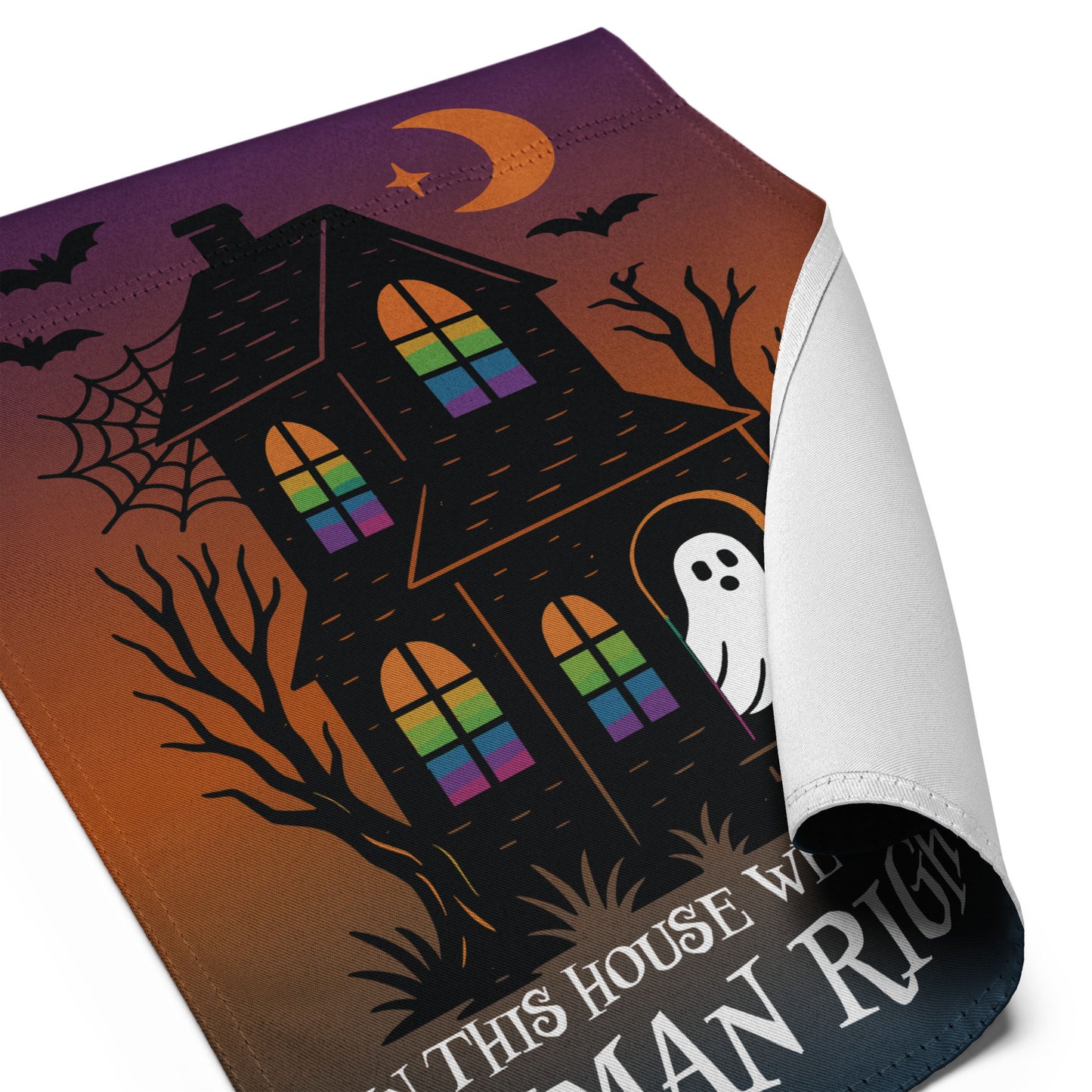 Angled close-up of the “In This House We Believe in Human Rights” Halloween garden flag with curled corner showing haunted house and spooky details, Blue Wave USA progressive products.