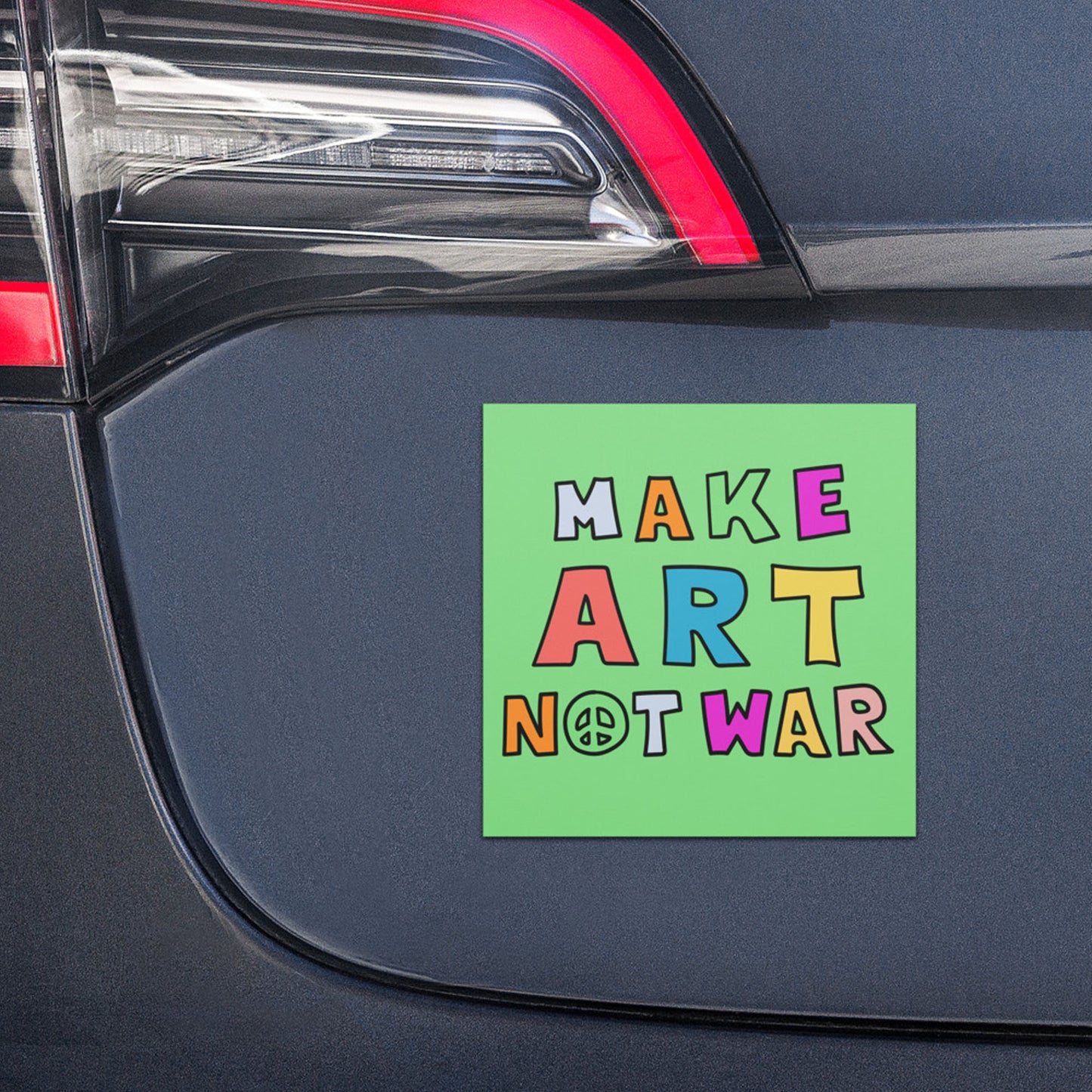 Green car magnet with multicolor “MAKE ART NOT WAR” typography and peace symbol displayed on vehicle trunk, creative anti-war message.