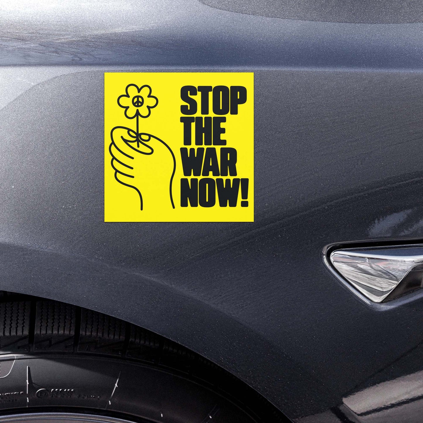 Yellow anti-war car magnet with hand-and-flower illustration and “STOP THE WAR NOW!” protest wording displayed on vehicle door panel.