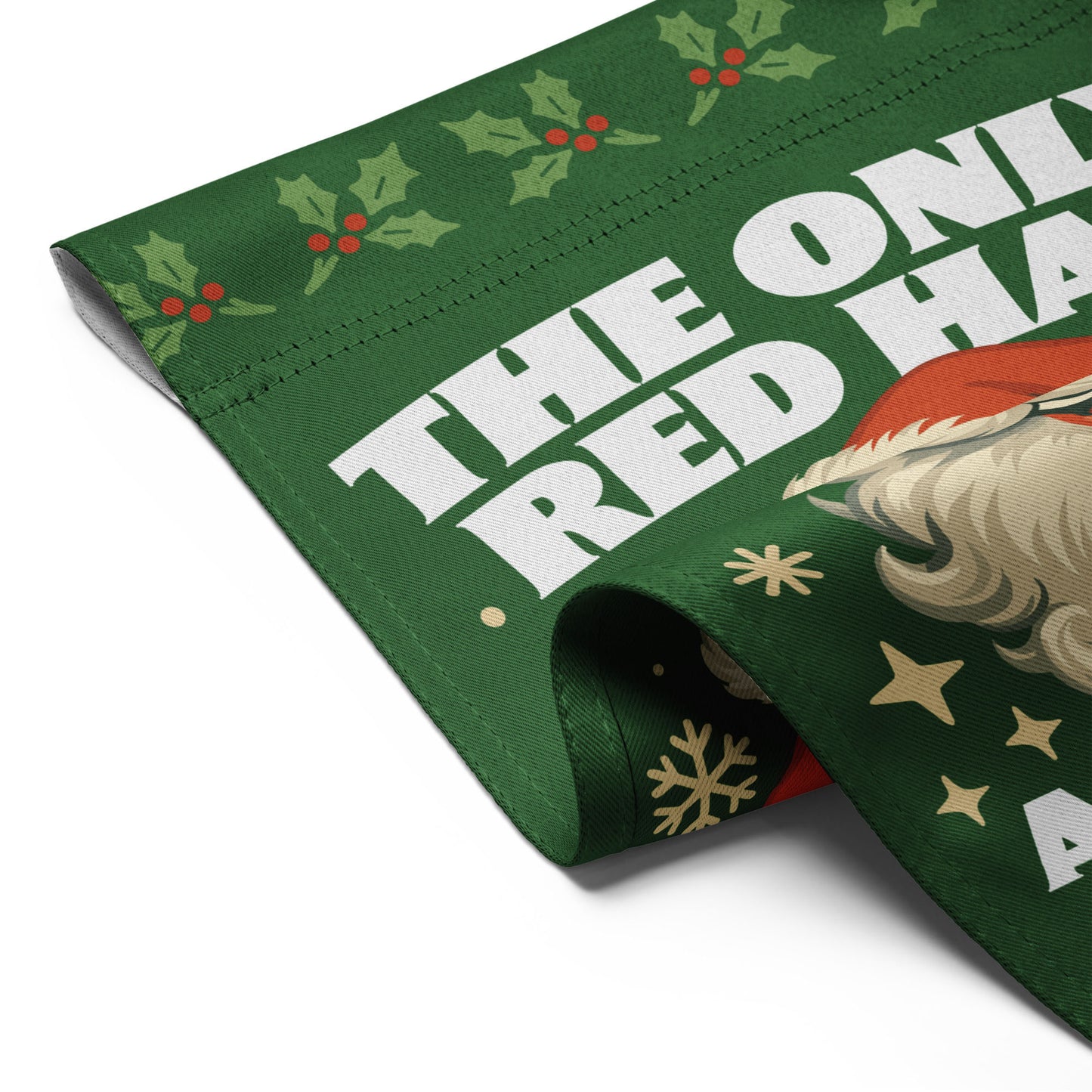 Close-up of a folded green garden flag featuring part of a retro Santa illustration, holiday accents, and the printed message “THE ONLY RED HAT ALLOWED IN OUR HOME.”