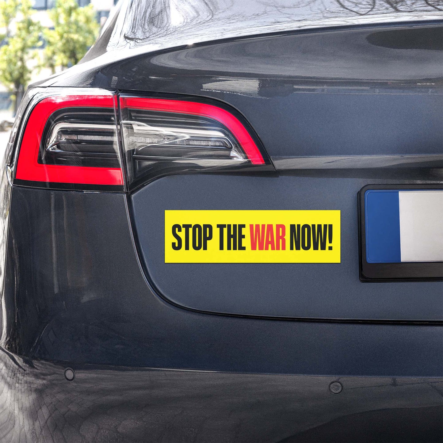 Yellow horizontal bumper magnet with “STOP THE WAR NOW!” wording displayed on rear of vehicle, high-contrast anti-war statement.
