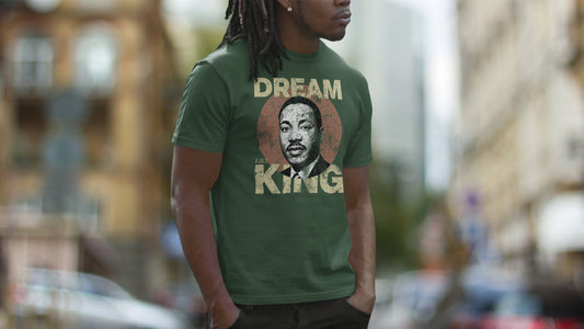 A man with dreadlocks wearing a dark green "Dream Like King" t-shirt stands outdoors in an urban setting.