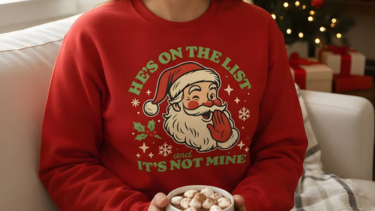 A person wearing a red Christmas sweatshirt with the playful vintage Santa and “He’s On the List & It’s Not Mine” text, styled for a cozy holiday moment. A popular pick from Blue Wave USA.