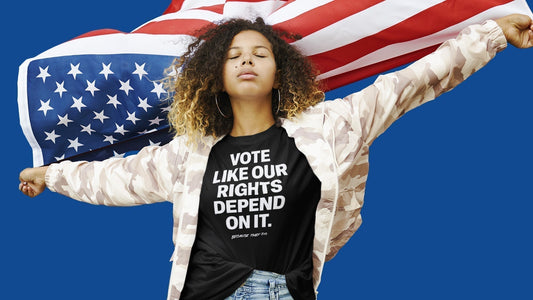 Person wearing a black “Vote Like Our Rights Depend On It. Because They Do.” t-shirt stands confidently with arms outstretched, holding an American flag against a bright blue background.