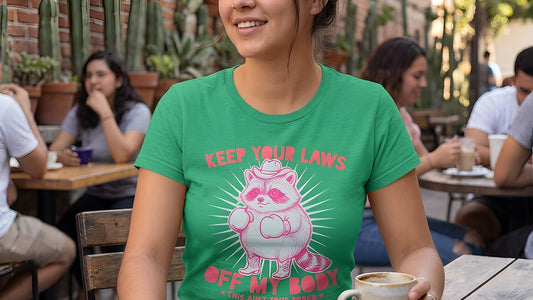Woman wearing green t-shirt with pink cowboy raccoon graphic and “Keep Your Laws Off My Body” text, styled in bold western feminist protest design.