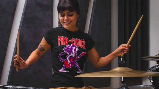 Woman playing drums while wearing black pro-choice graphic t-shirt featuring punk rock cat with electric guitar and bold “PRO-CHOICE” feminist design.