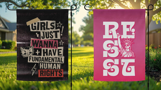 Two progressive political garden flags displayed side-by-side in a green yard: a black punk flag reading 'Girls Just Wanna Have Fundamental Human Rights' and a pink retro flag reading 'Resist'.