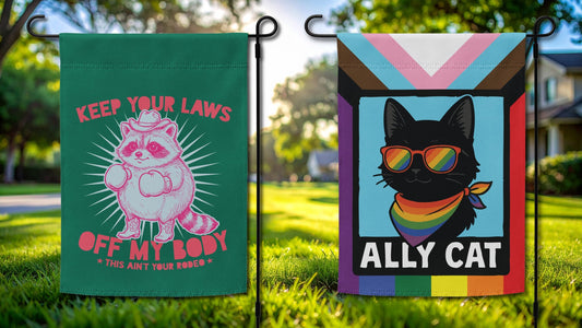 Two progressive garden flags in a sunny yard: a green "Keep Your Laws Off My Body" cowboy raccoon flag and a rainbow "Ally Cat" LGBTQ+ Pride flag.