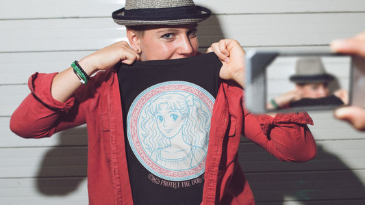 Person in a red shirt and fedora posing with a black "PROTECT THE DOLLS" t-shirt featuring retro anime graphic design.
