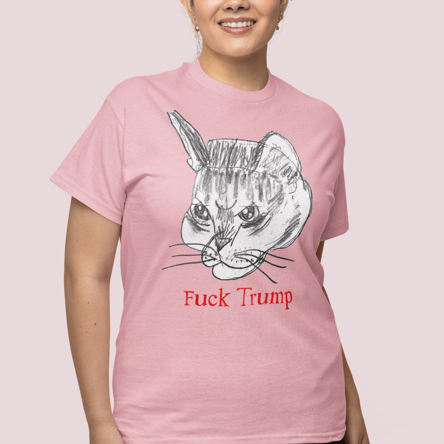 Model wearing light pink unisex t-shirt featuring illustrated cat head graphic with red text reading “Fuck Trump,” relaxed fit.