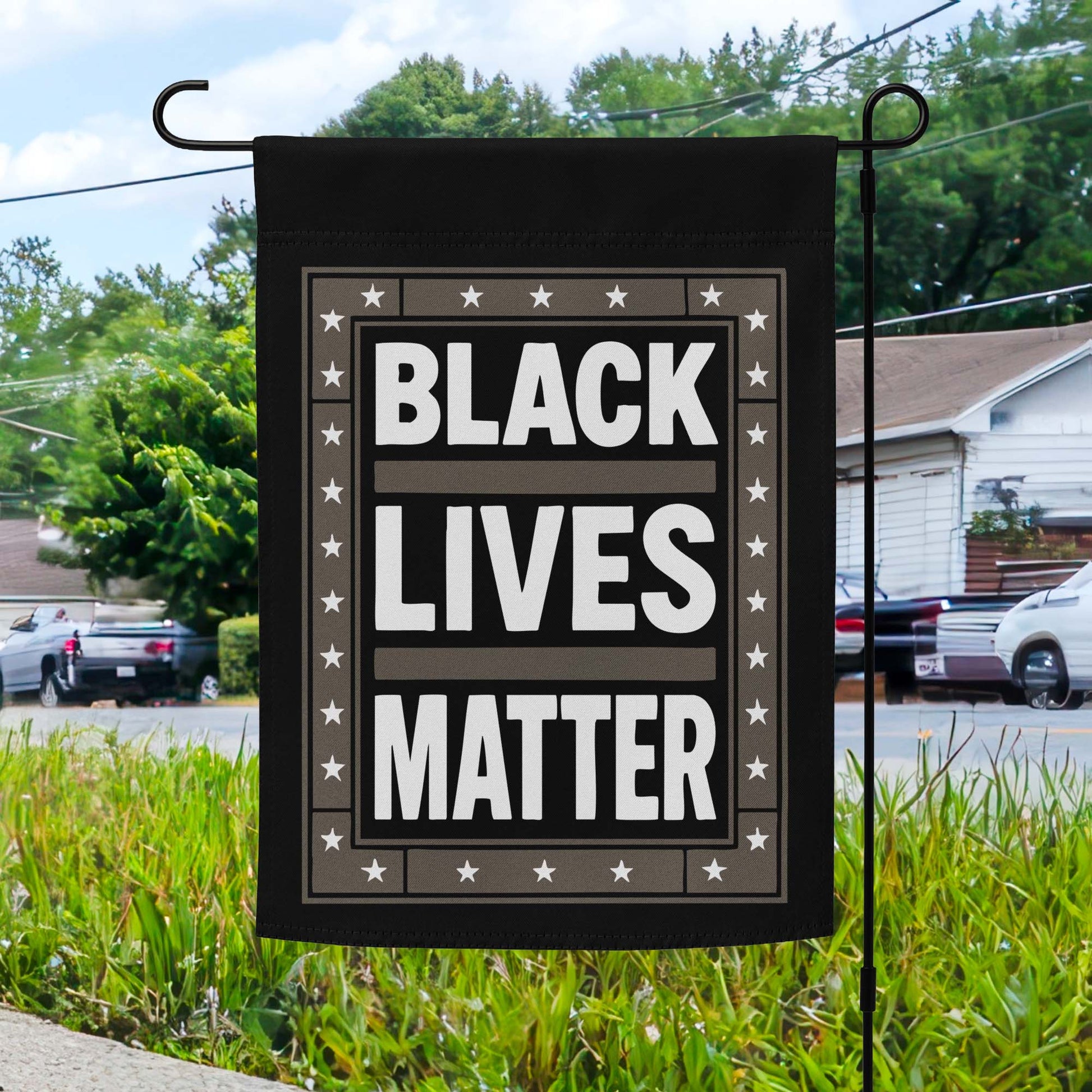 Black Lives Matter garden flag with bold white text on black background and star border, displayed outdoors on a flagpole in front of a home.