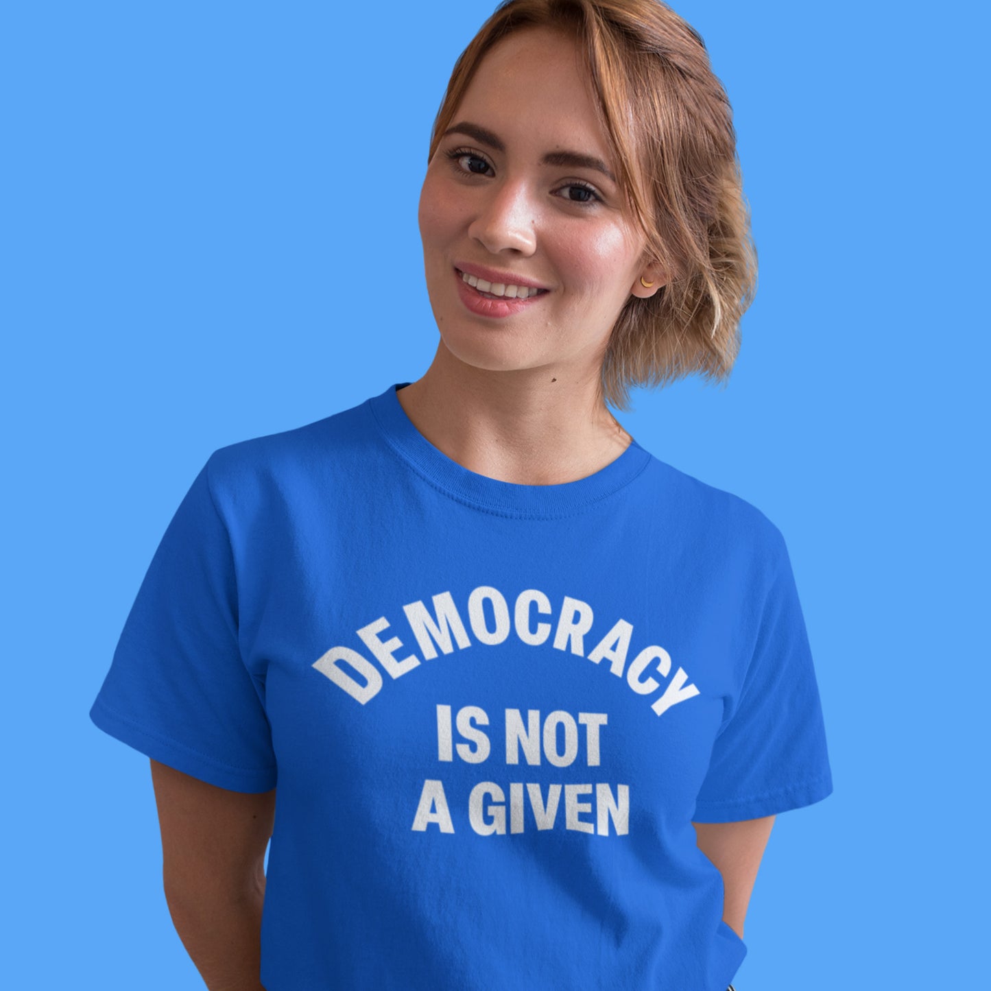 Model wearing blue unisex classic tee featuring bold white text reading “DEMOCRACY IS NOT A GIVEN.”