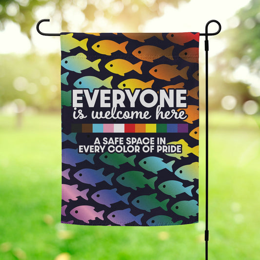 “Everyone Is Welcome Here – A Safe Space In Every Color of Pride” garden flag with rainbow fish pattern, displayed outdoors on a flagpole.