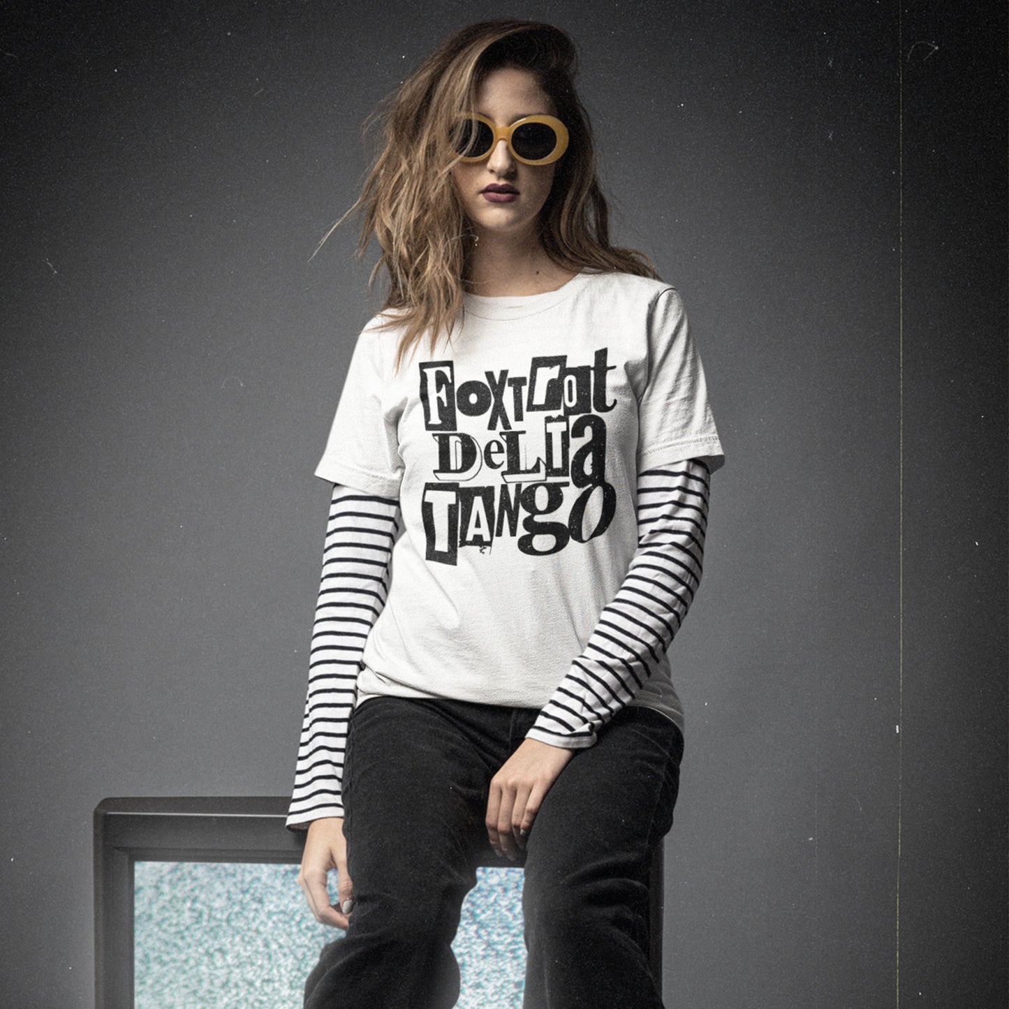 Model wearing white unisex t-shirt with stacked block text reading “Foxtrot Delta Tango,” high-contrast black lettering, casual studio styling.