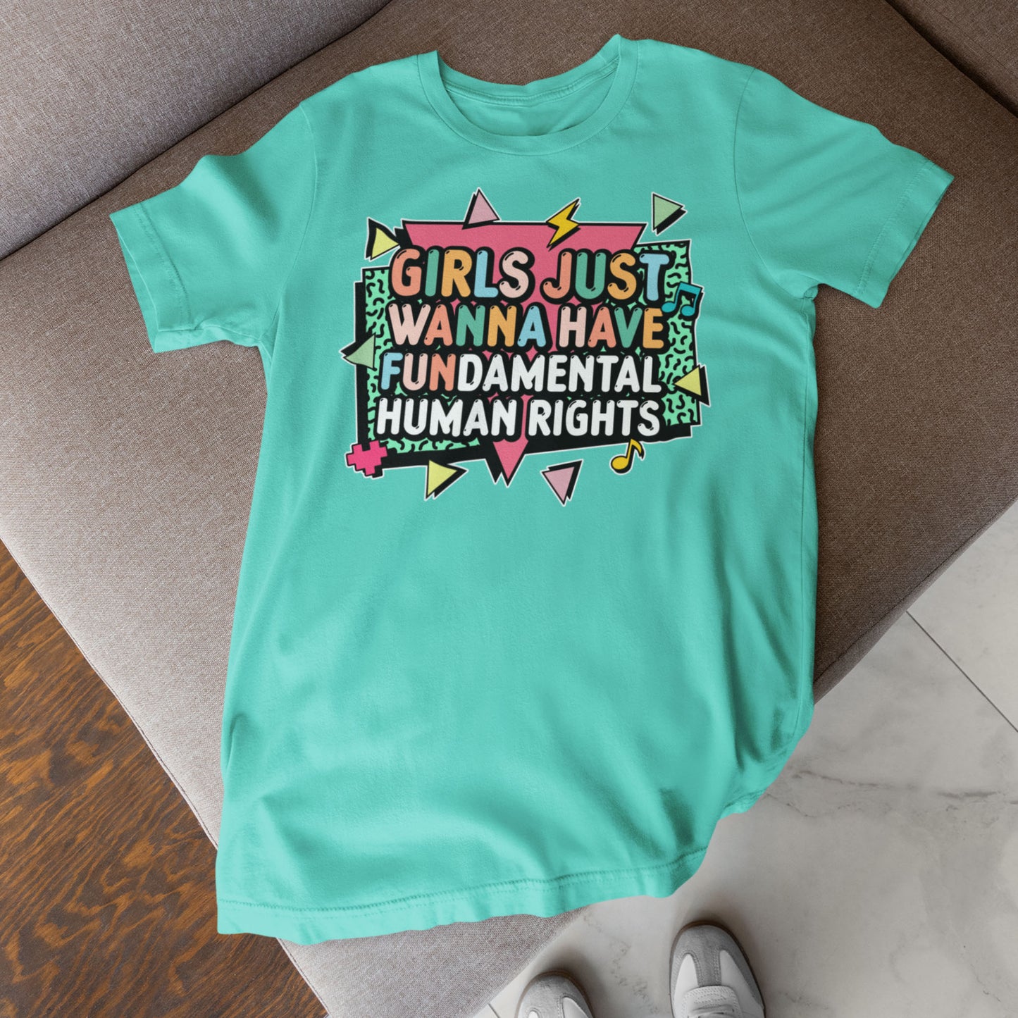 Turquoise t-shirt laid flat on a sofa with bold retro lettering that reads “Girls Just Wanna Have Fundamental Human Rights,” surrounded by pastel lightning bolts and playful geometric shapes. Empowering feminist tee by Blue Wave USA.