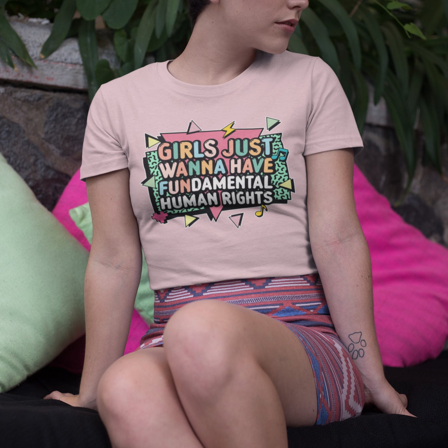 Model sitting outdoors wearing a light pink “Girls Just Wanna Have Fundamental Human Rights” tee with 80s-style lightning bolt design. Empowering women’s rights shirt by Blue Wave USA.