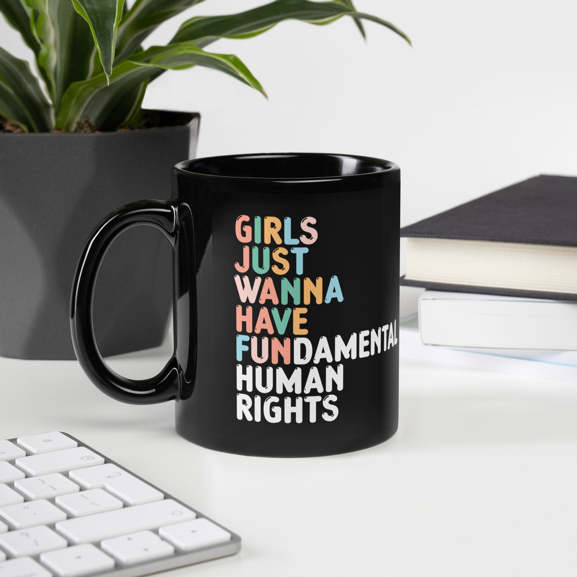 Black feminist coffee mug with “Girls Just Wanna Have Fundamental Human Rights” slogan displayed beside keyboard and indoor plant workspace setup.