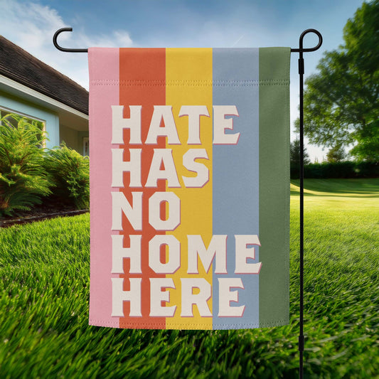 Progressive garden flag featuring retro stacked typography reading “HATE HAS NO HOME HERE,” vertical rainbow stripe background, displayed outdoors on metal stand.