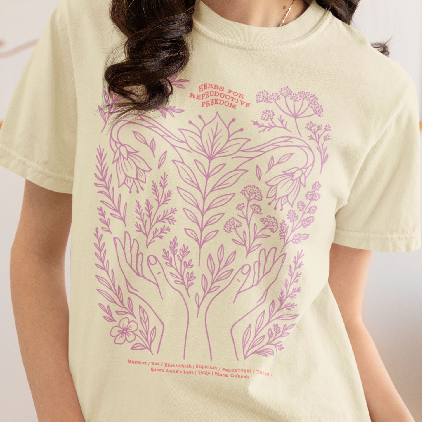 Model wearing the cream “Herbs for Reproductive Freedom” unisex tee showing delicate pink floral illustration inspired by traditional herbal medicine and body autonomy.