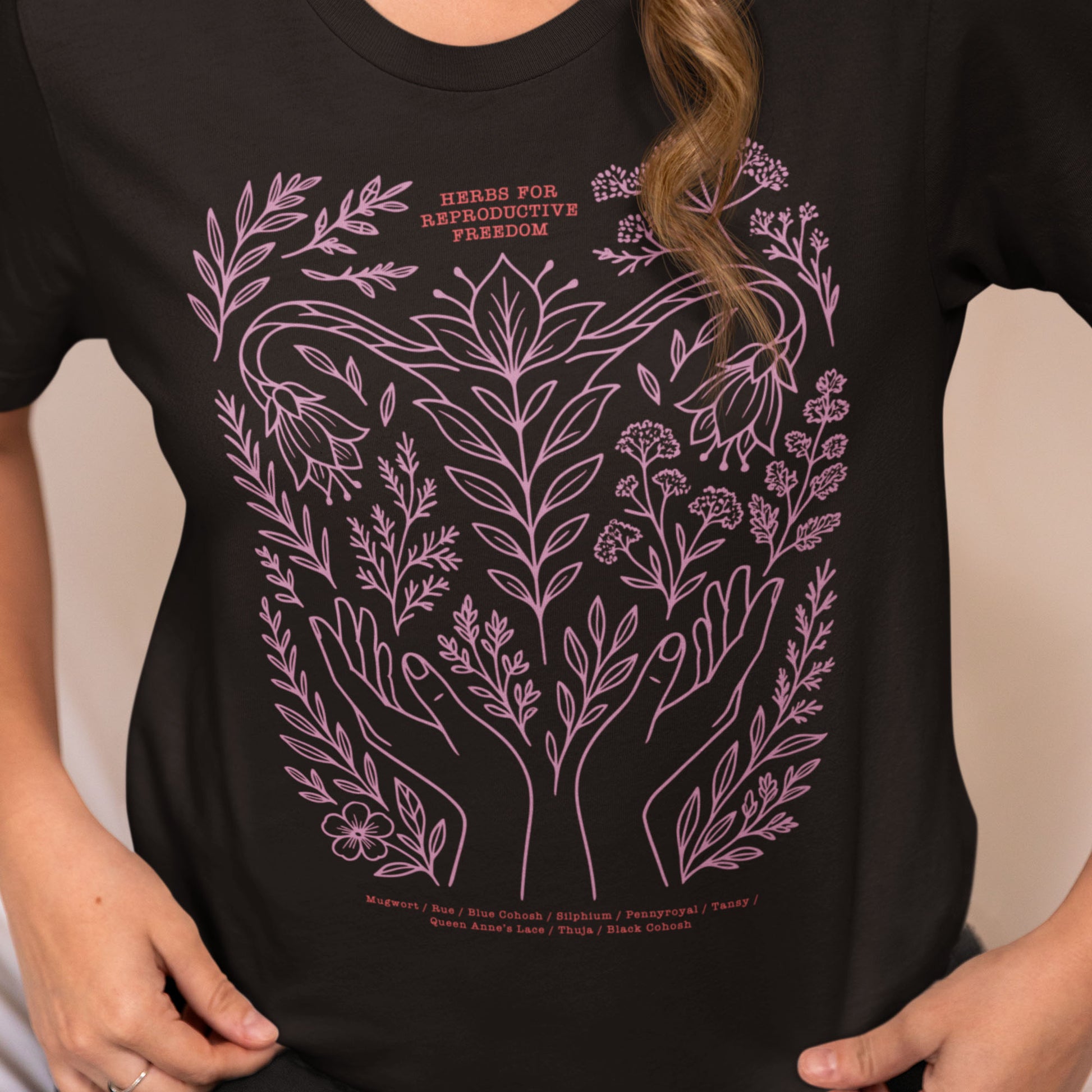 Model wearing the black “Herbs for Reproductive Freedom” unisex t-shirt featuring the lavender design of healing herbs like mugwort, rue, and blue cohosh. Feminist botanical tee by Blue Wave USA.