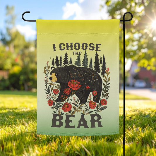 Light green garden flag displayed outdoors on a metal stand, featuring a folk-art style bear surrounded by flowers and trees with bold text reading “I CHOOSE THE BEAR.”