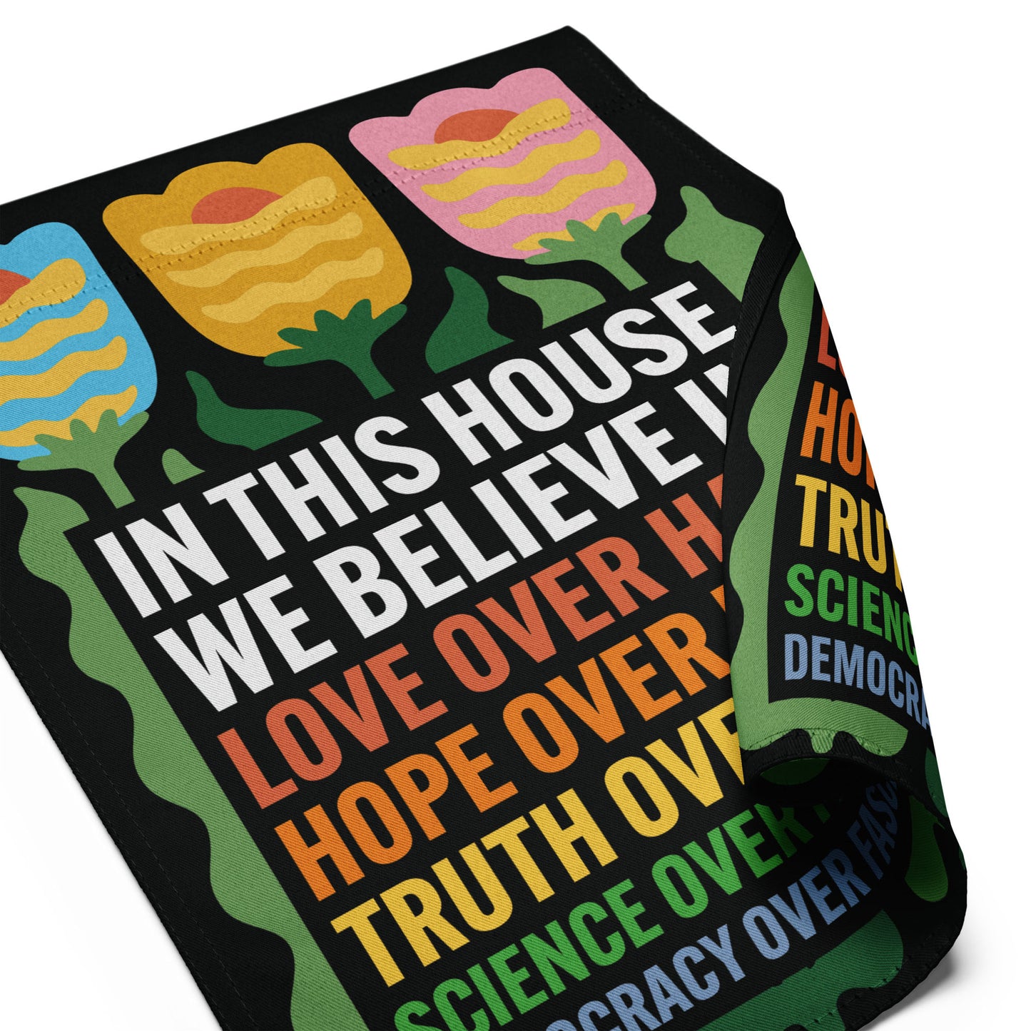 Close-up of black garden flag fabric showing stitched edges, folk-art tulip details, and multicolor “In This House We Believe” lettering.