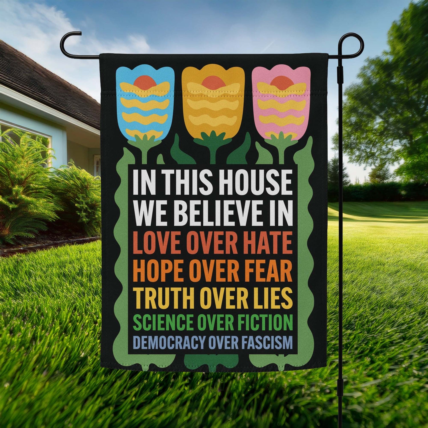 Black garden flag featuring folk-art tulip illustrations and stacked multicolor text reading “IN THIS HOUSE WE BELIEVE IN LOVE OVER HATE HOPE OVER FEAR TRUTH OVER LIES SCIENCE OVER FICTION DEMOCRACY OVER FASCISM.”