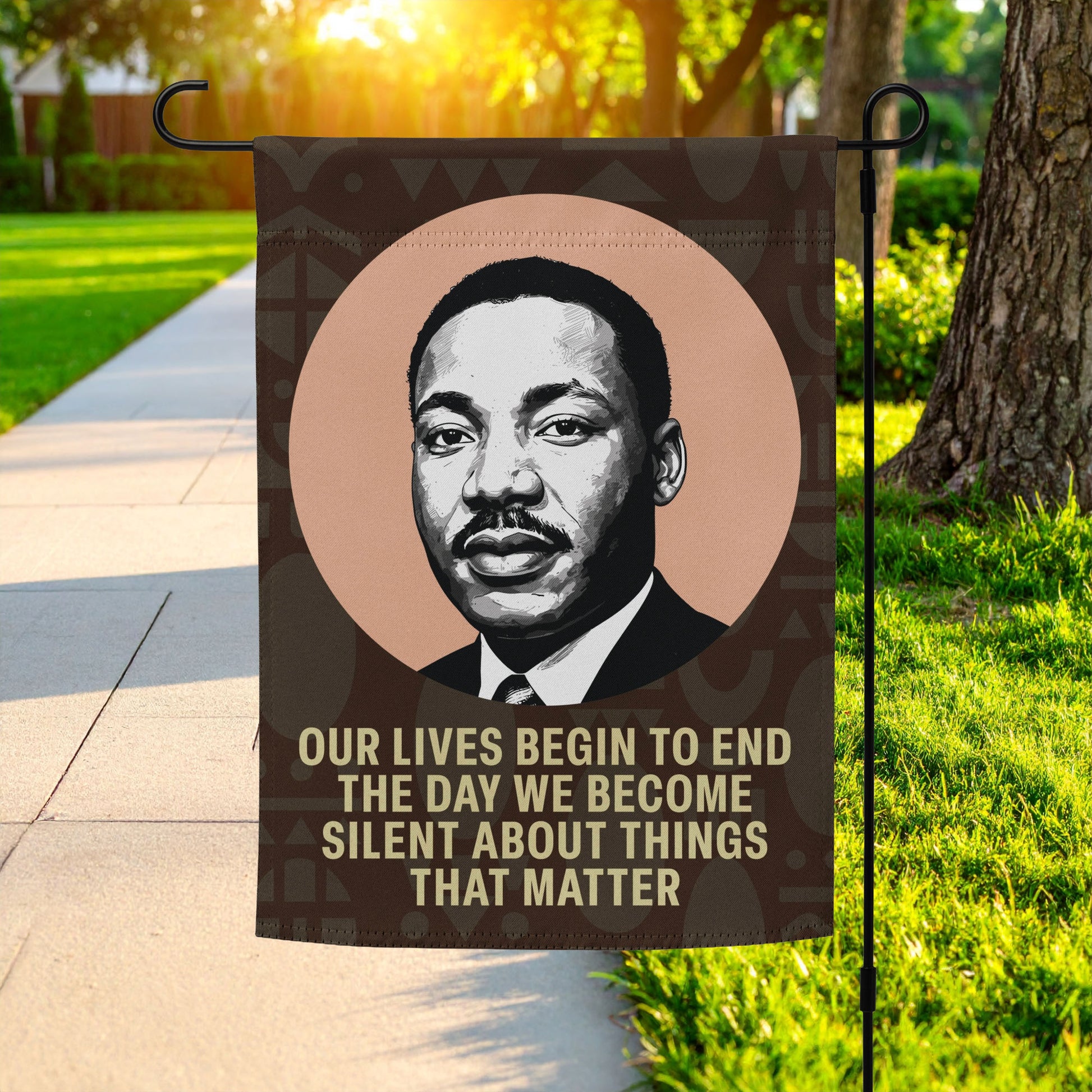 A garden flag printed with Martin Luther King Jr.’s portrait and the quote “OUR LIVES BEGIN TO END THE DAY WE BECOME SILENT ABOUT THINGS THAT MATTER,” displayed outdoors along a sunny walkway.