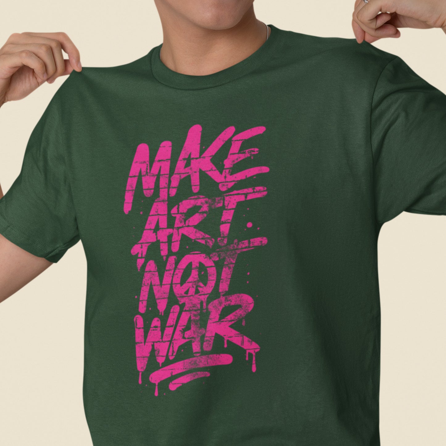 Model wearing a forest green unisex t-shirt with bright pink brush-style typography spelling “MAKE ART NOT WAR,” centered graphic shown in a casual studio setting.