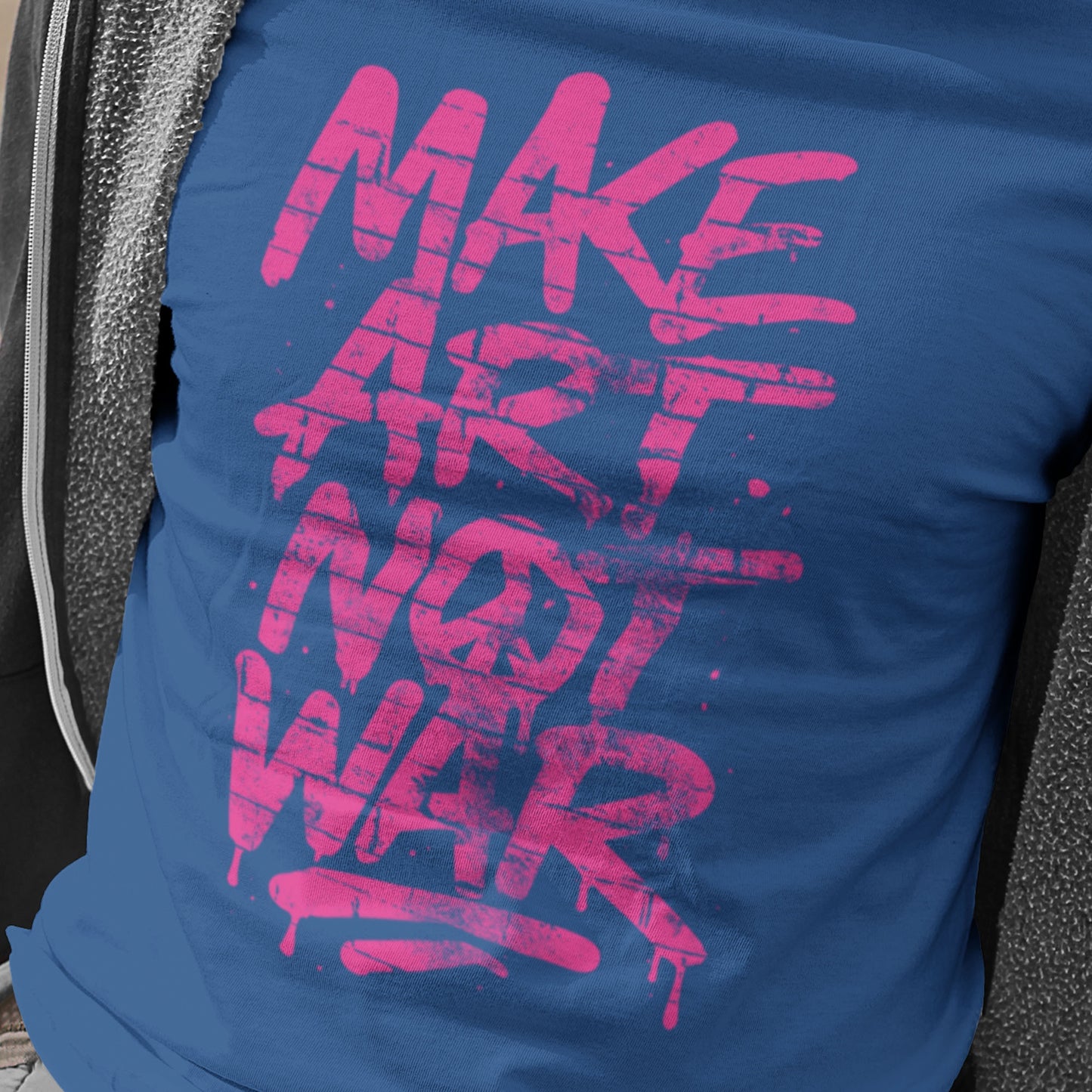 Close-up of blue unisex t-shirt featuring graffiti-style artwork with dripping pink text reading “MAKE ART NOT WAR.”, street-art inspired activist design.