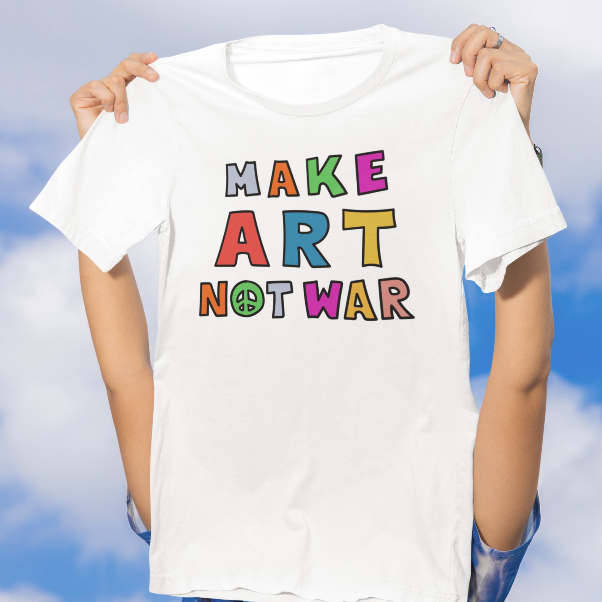 Model holding a white unisex t-shirt against a sky backdrop, displaying colorful block-style lettering spelling “MAKE ART NOT WAR,” centered graphic with a playful activist tone from Blue Wave USA.