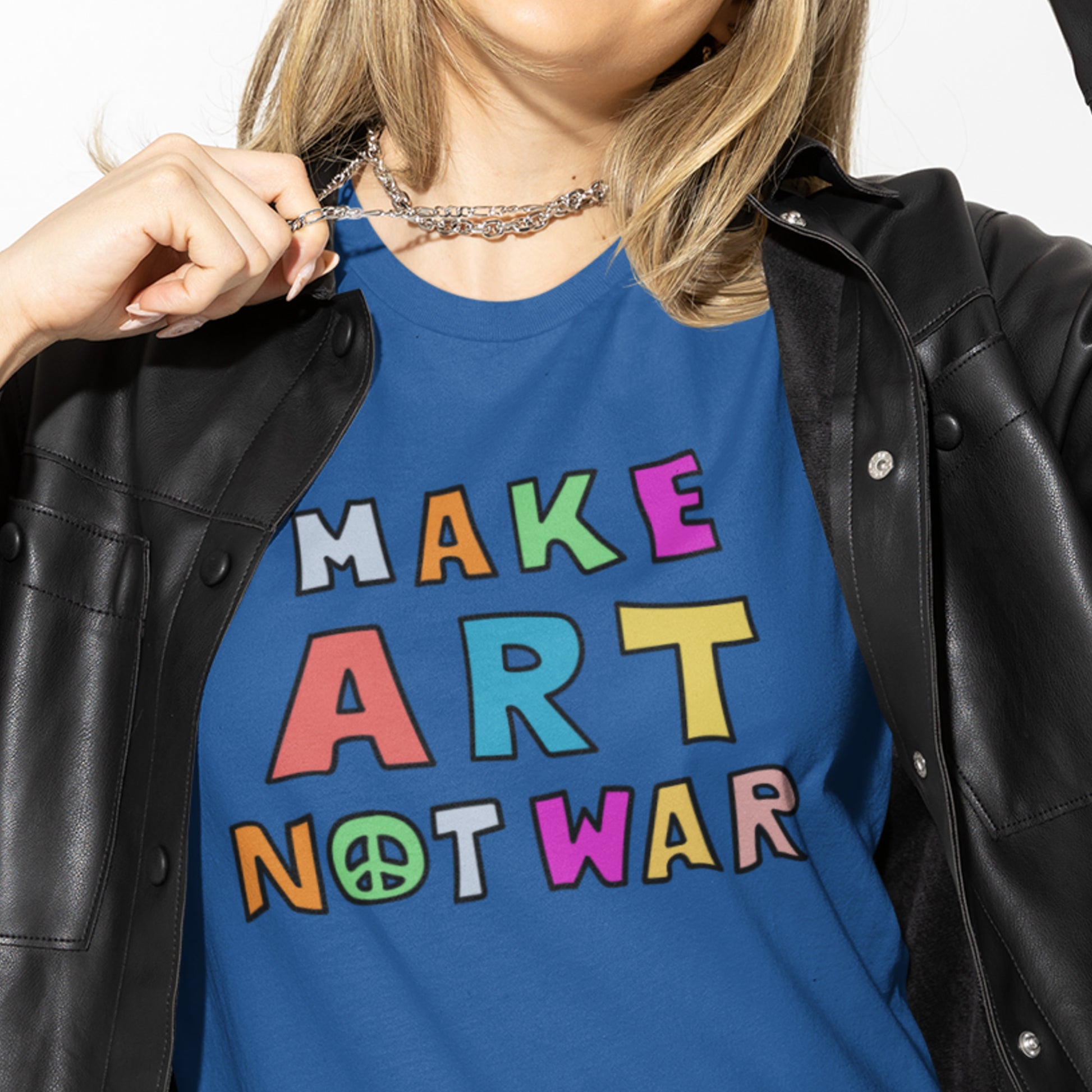 Model wearing a blue unisex t-shirt layered under a jacket, showing multicolor block lettering reading “MAKE ART NOT WAR,” casual lifestyle styling in a studio setting.
