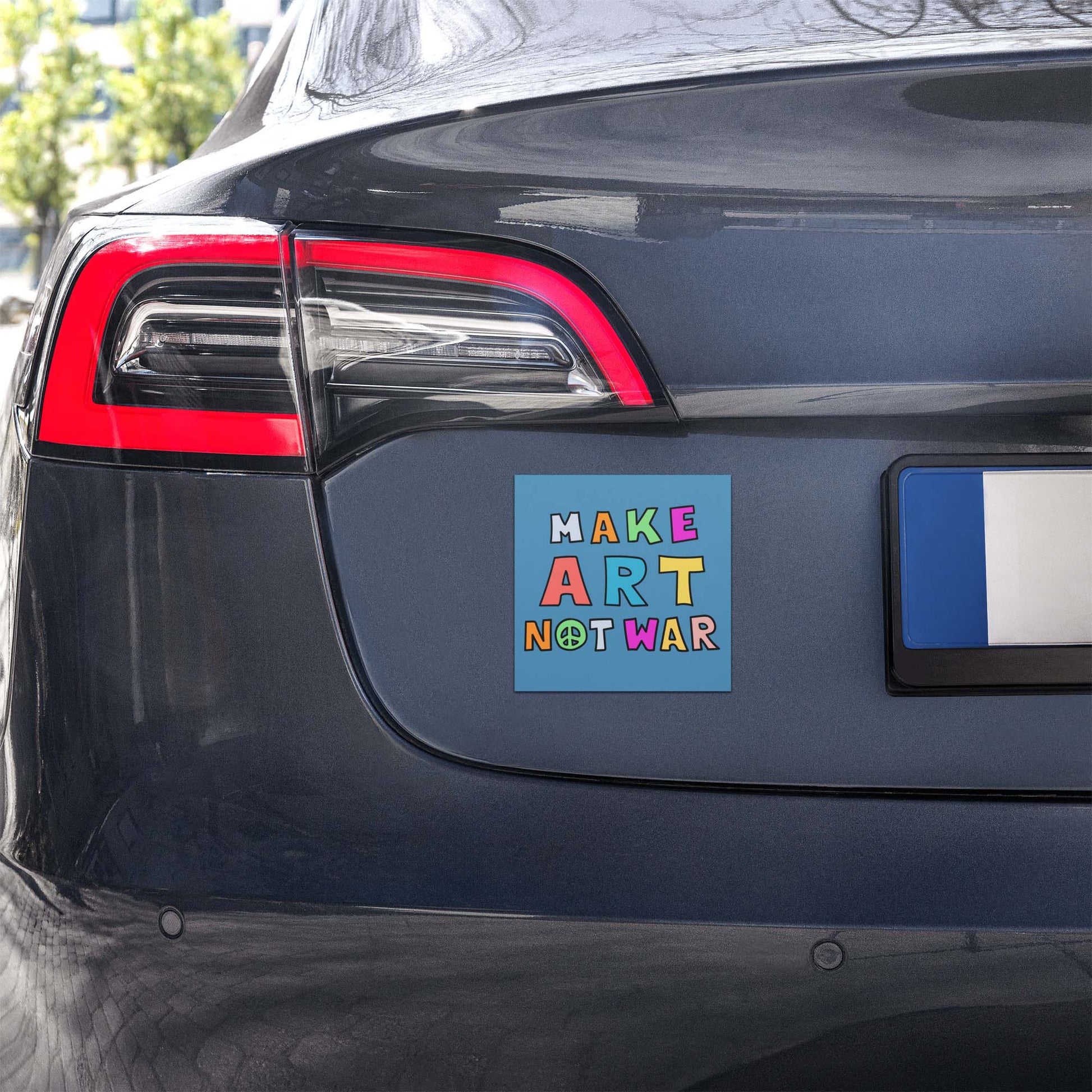 Blue square car magnet displayed on a vehicle bumper outdoors, showing multicolor block lettering with peace symbol reading “MAKE ART NOT WAR,” anti-war magnet from Blue Wave USA.