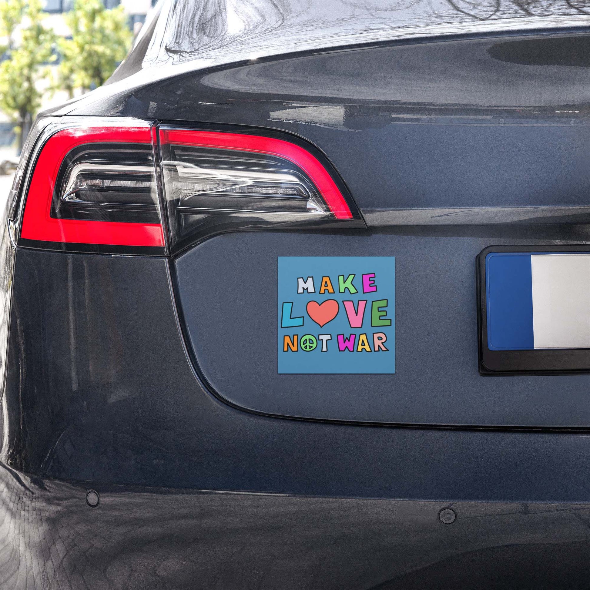 Blue square car magnet displayed on a vehicle bumper outdoors, showing multicolor block lettering with heart and peace symbol reading “MAKE LOVE NOT WAR,” anti-war magnet from Blue Wave USA.